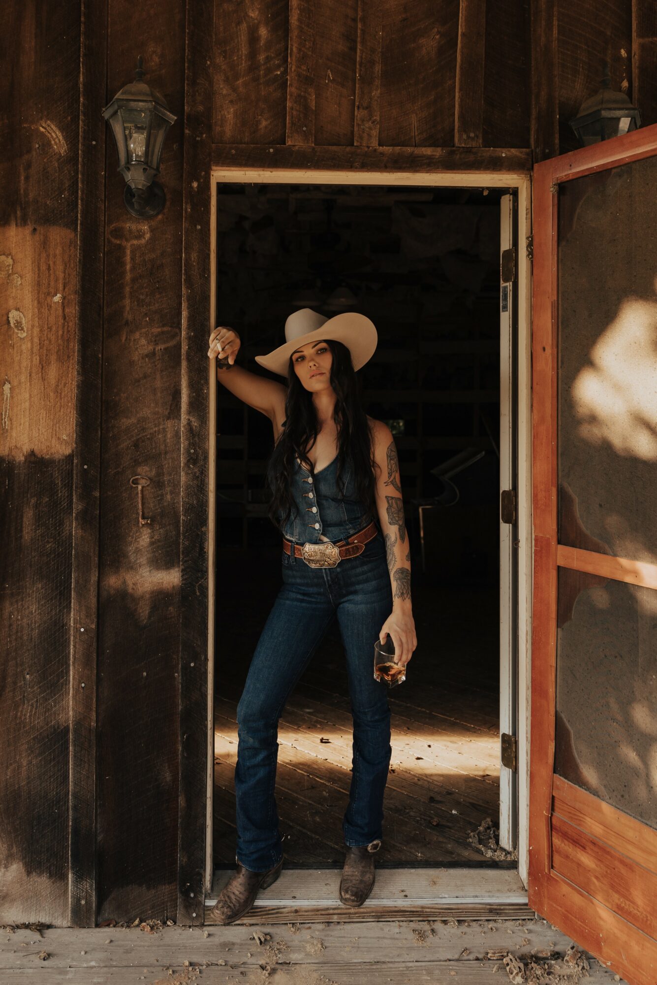 Woman in a hat standing in doorway, wooden wall background, holding a drink, wearing jeans and a sleeveless top.