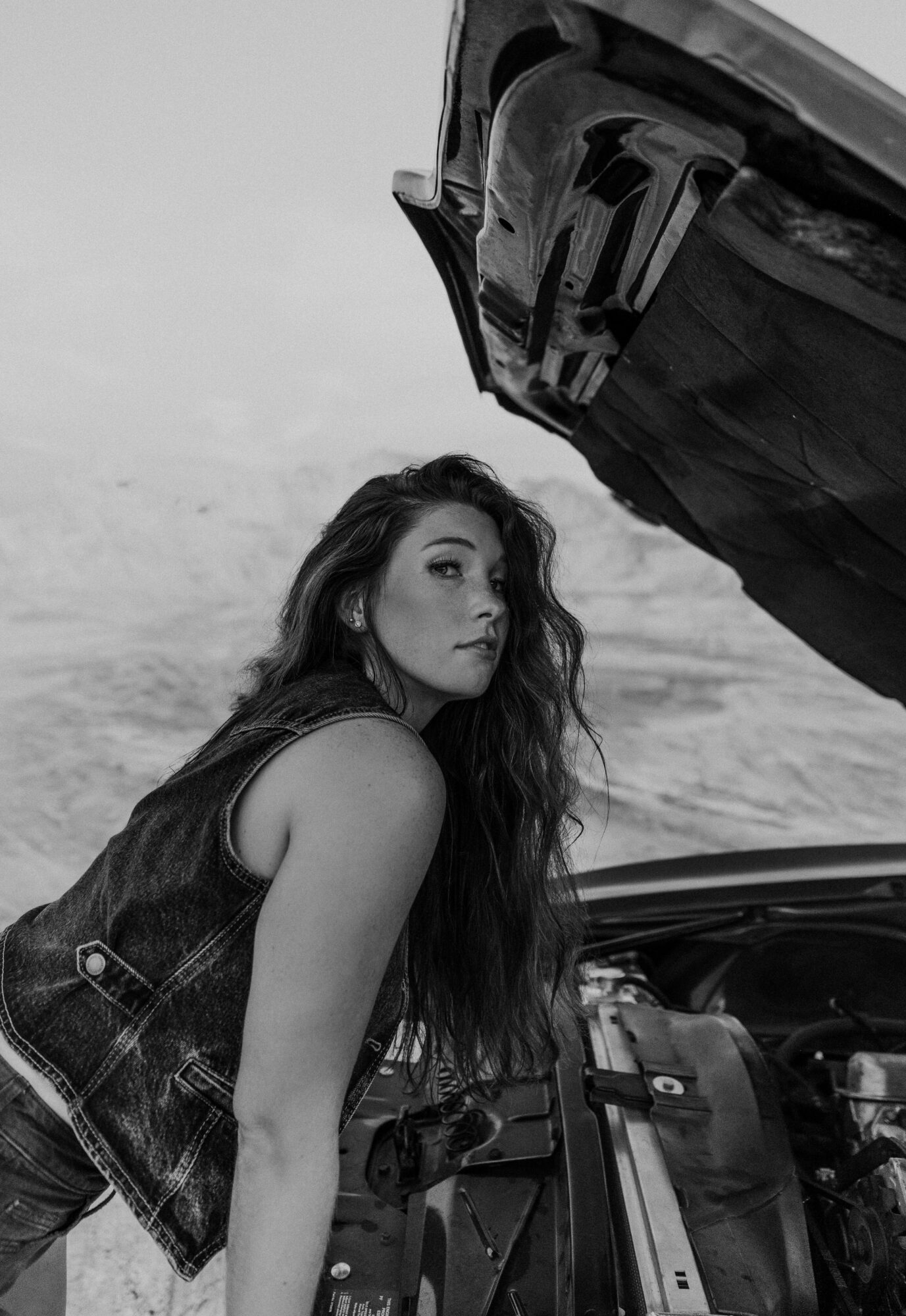 Woman with long hair leaning over open car hood outdoors, looking at camera, black and white image.