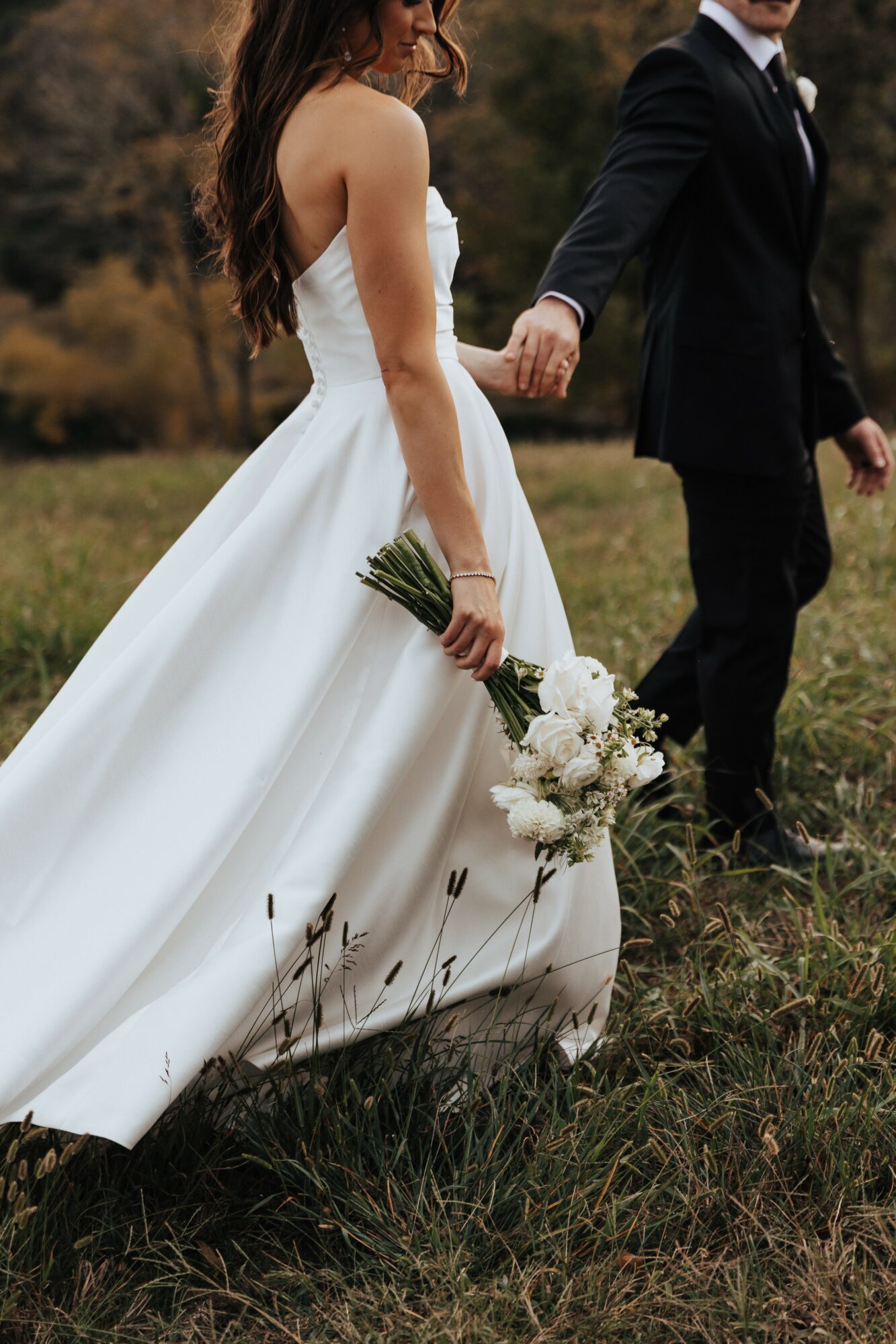 Bride in a white wedding dress holding a bouquet, walking with a groom in a black suit outdoors.