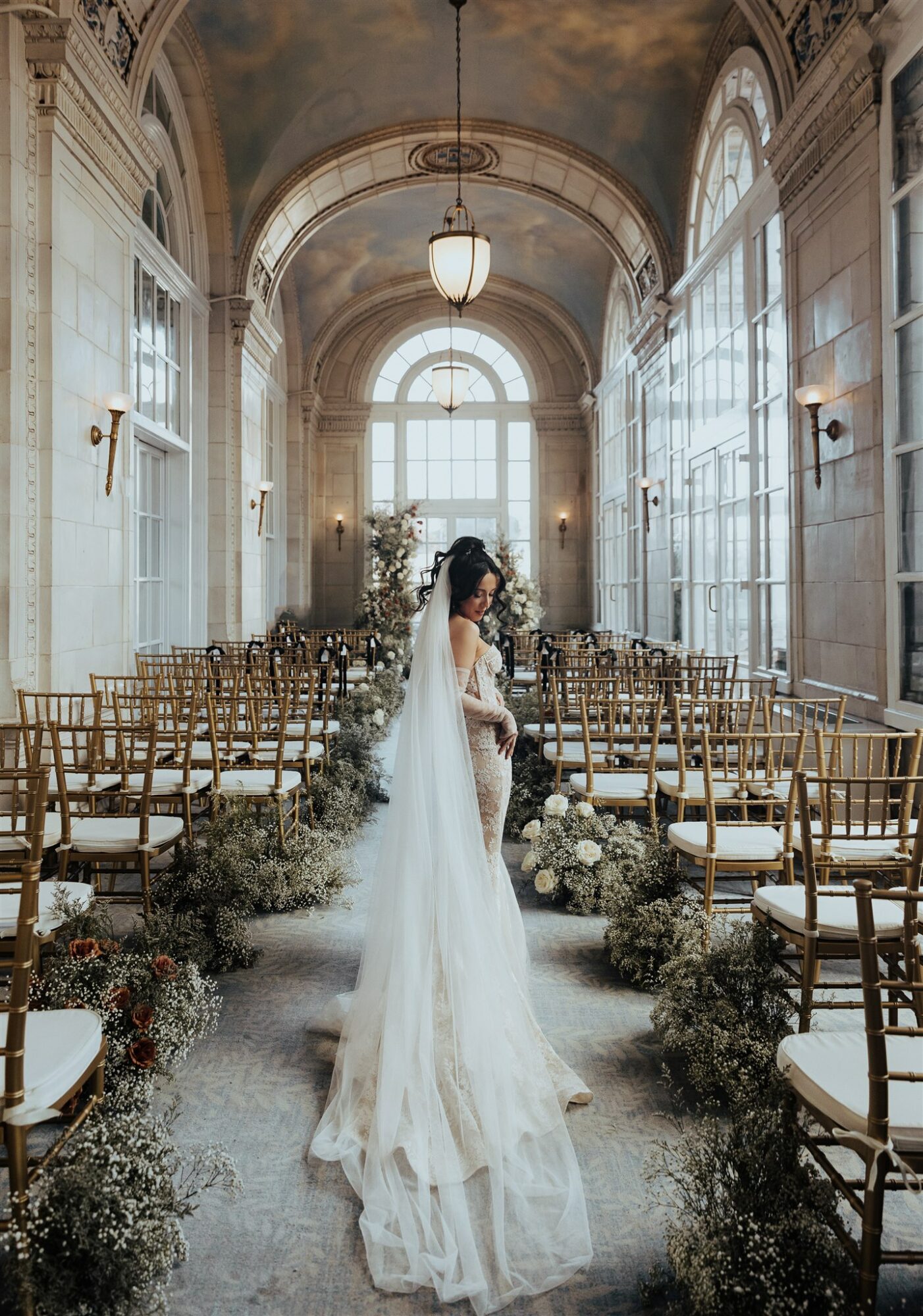 Bride in a wedding gown standing in a grand, decorated hall with large windows and rows of chairs.