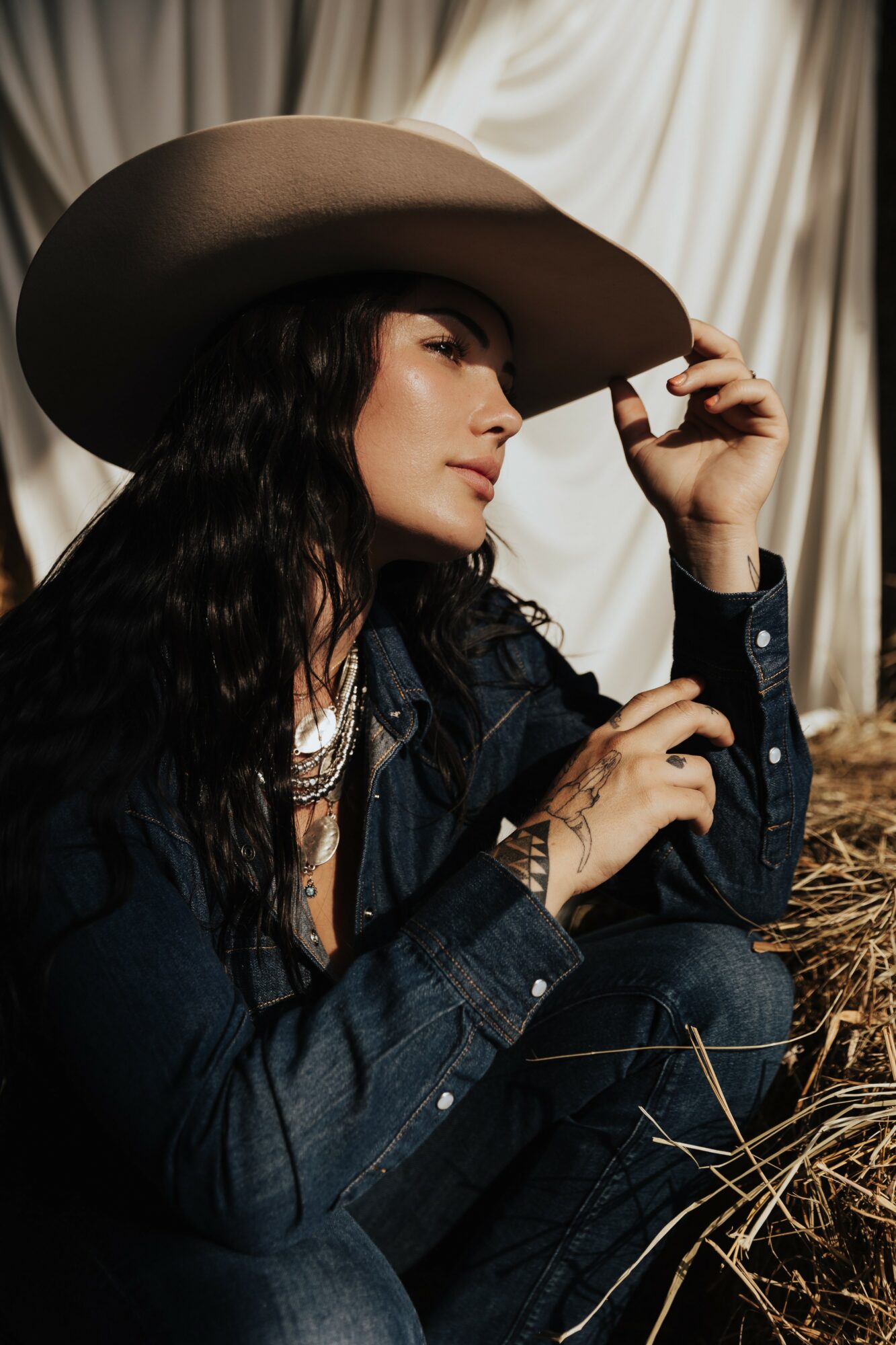 Woman wearing a large hat, sitting with a curtain backdrop, holding the brim of her hat, with long dark hair and denim clothing.