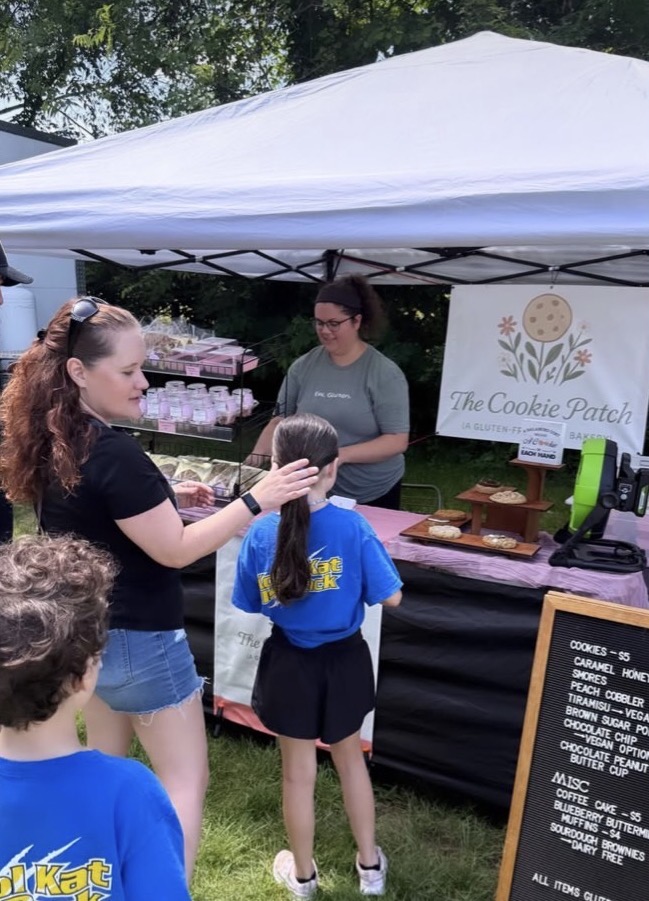 People at a cookie stand under a white canopy, with a sign reading 'The Cookie Patch,' outdoors with trees in background.