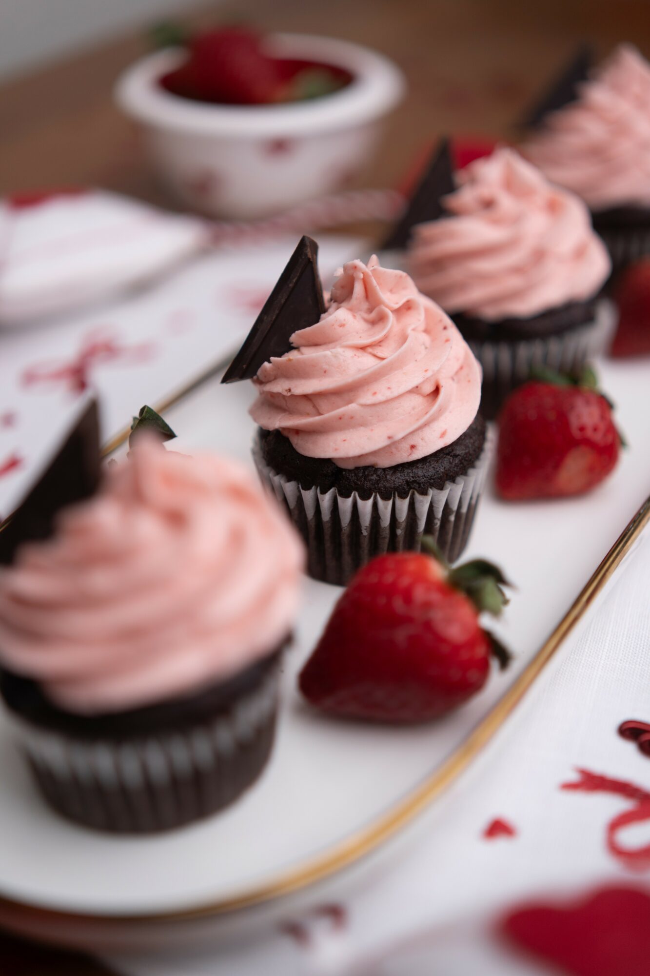 Chocolate cupcakes with pink frosting, strawberries, and a white plate with a decorative pattern, on a table.