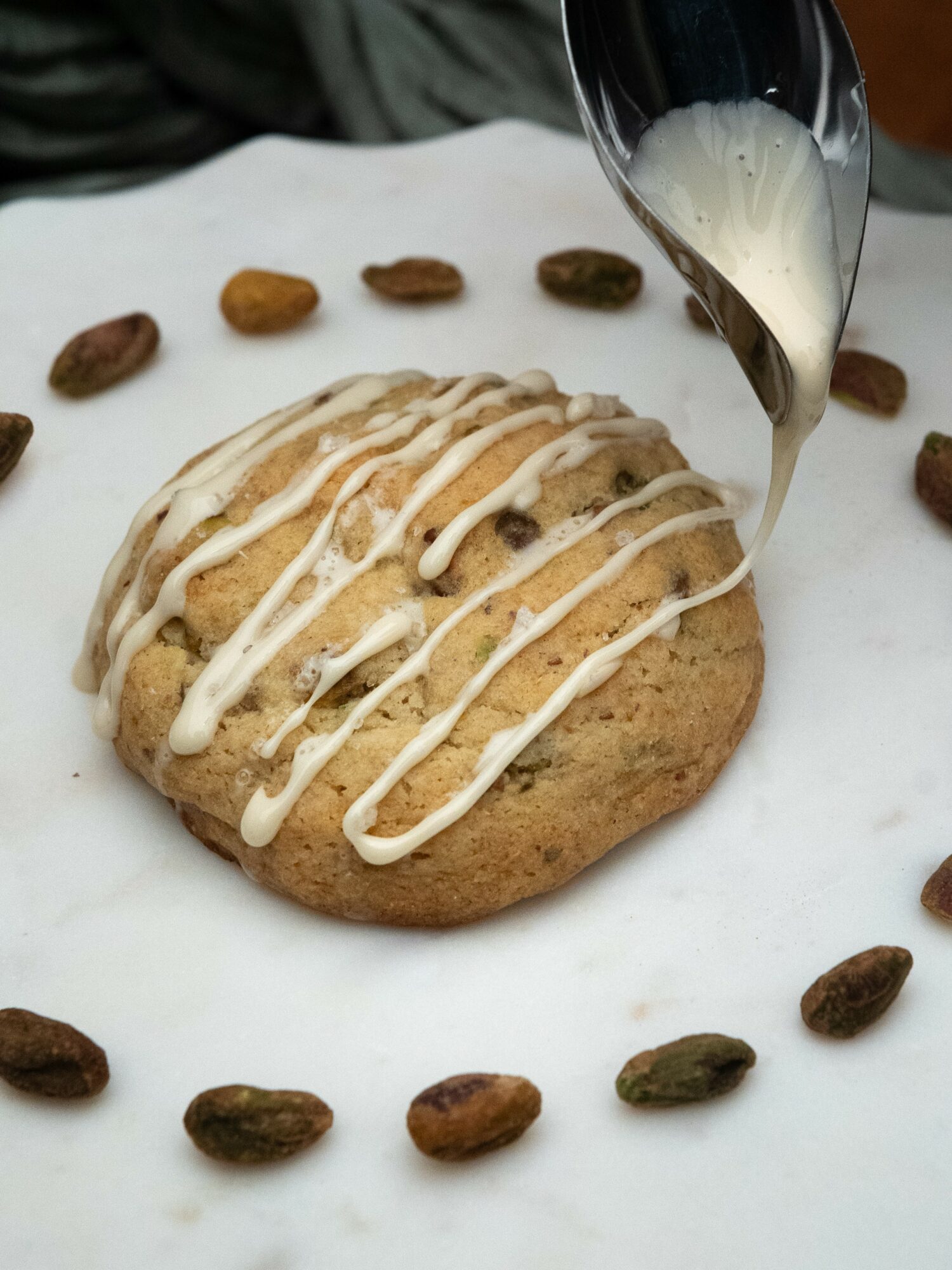 A cookie with white icing being decorated with white icing from a piping bag, surrounded by scattered pistachio nuts on white paper