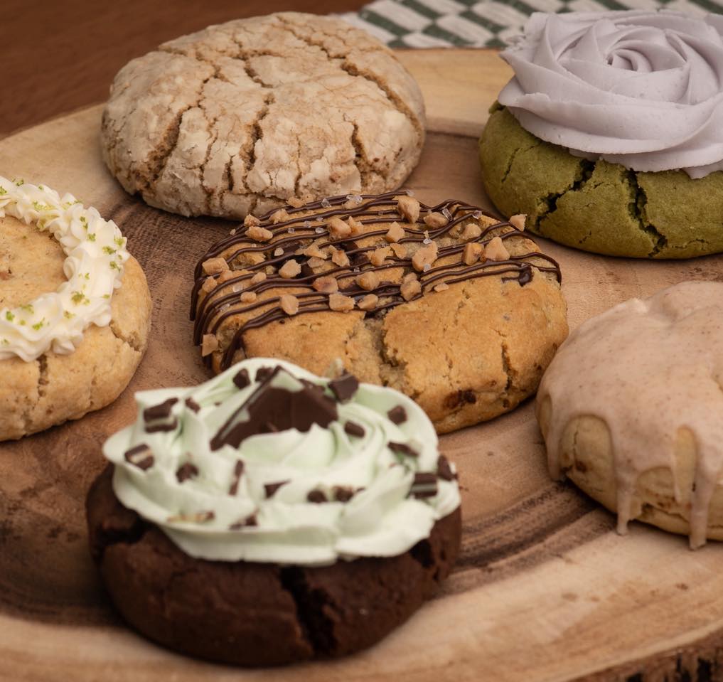Six cookies with different toppings on a wooden surface, including chocolate, icing, and a purple flower-shaped decoration.