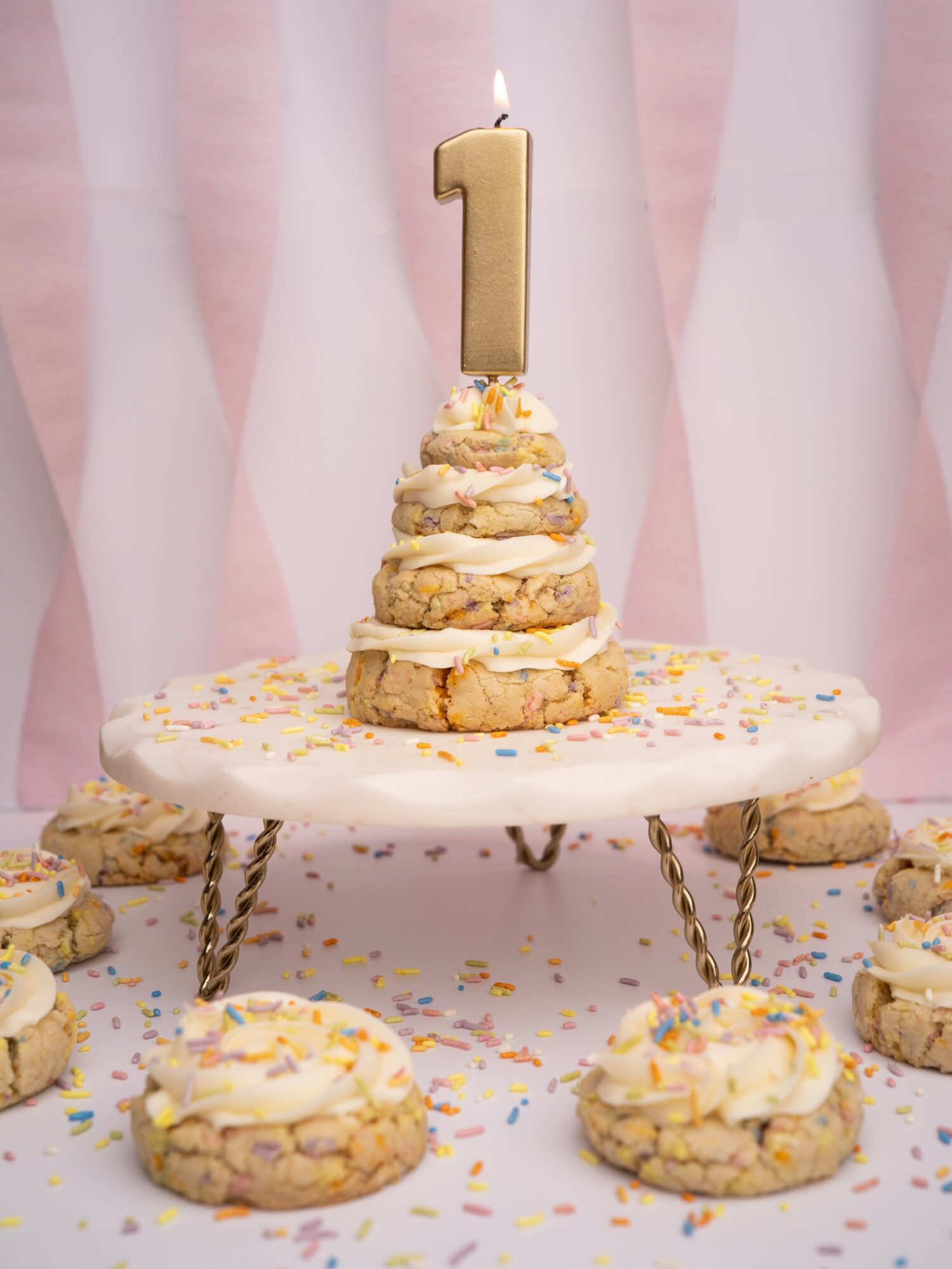 Cake with a large number one candle, decorated with sprinkles, on a table with cookies and colorful confetti, pink backdrop.