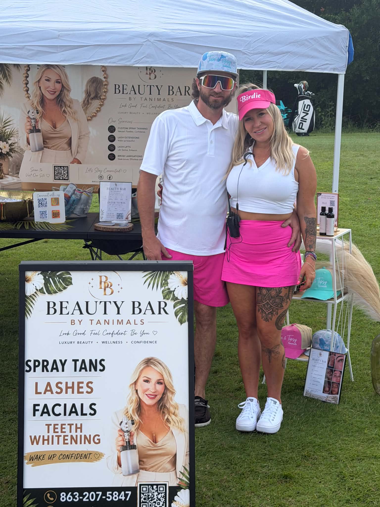 Two people stand in front of a beauty bar booth outdoors, smiling, with promotional signs and products displayed.