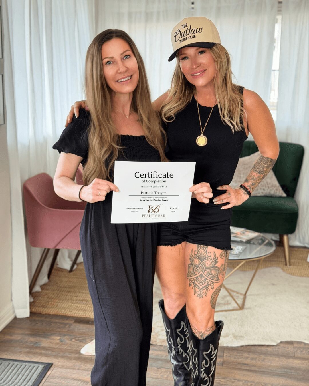 Two women smiling, one holding a certificate, standing indoors with curtains and furniture in background.