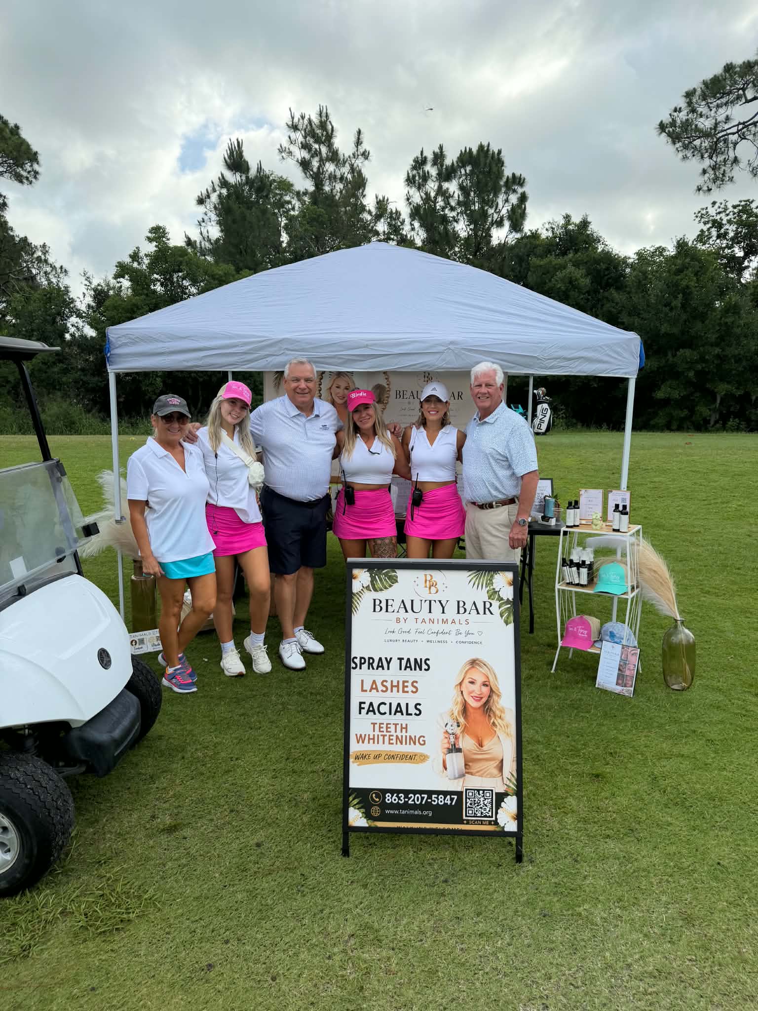 Group of six people standing under a white canopy tent outdoors, with a sign for a beauty bar and products nearby.