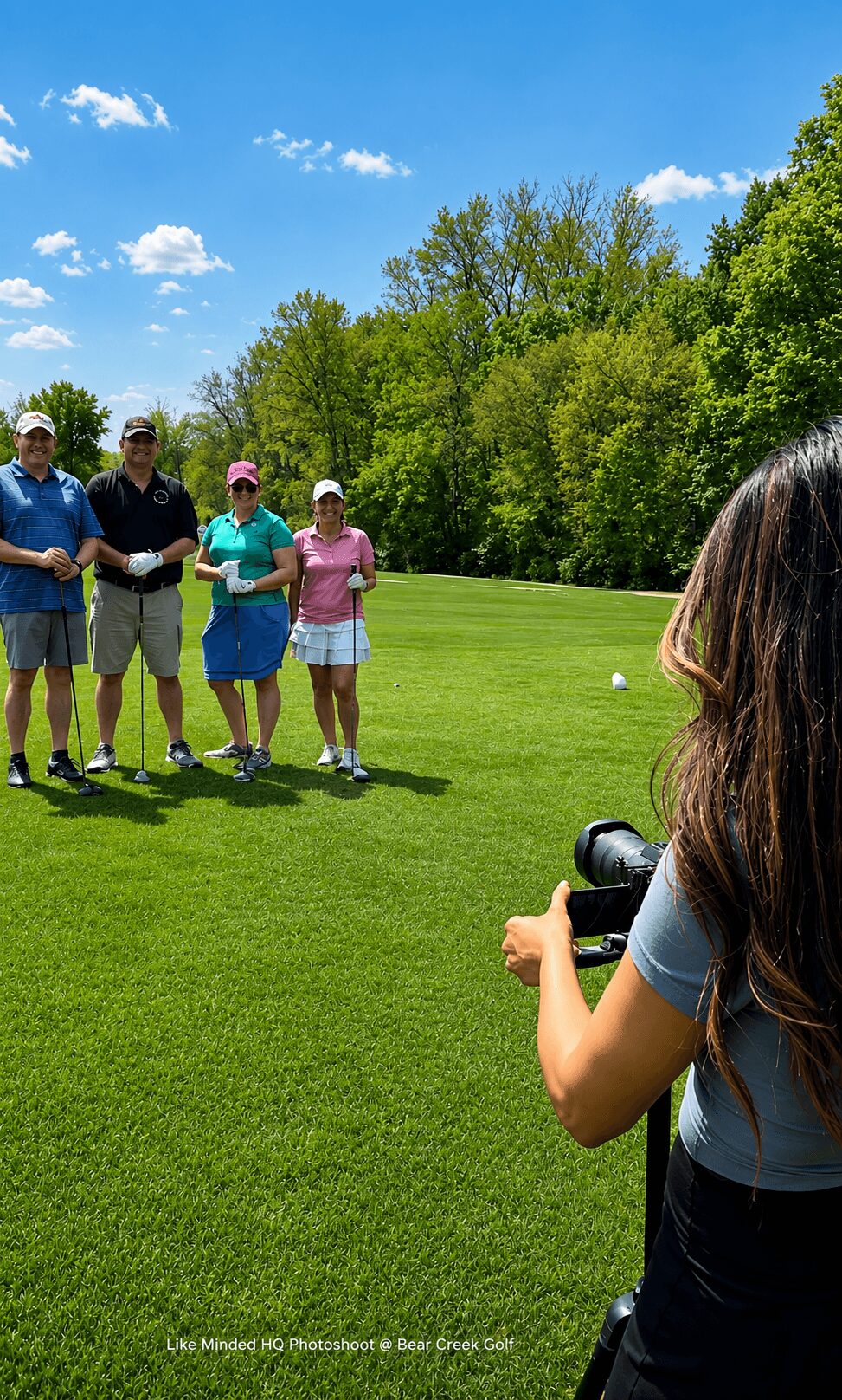 Four people standing on a golf course, with a woman taking a photo of them, trees and blue sky in background.