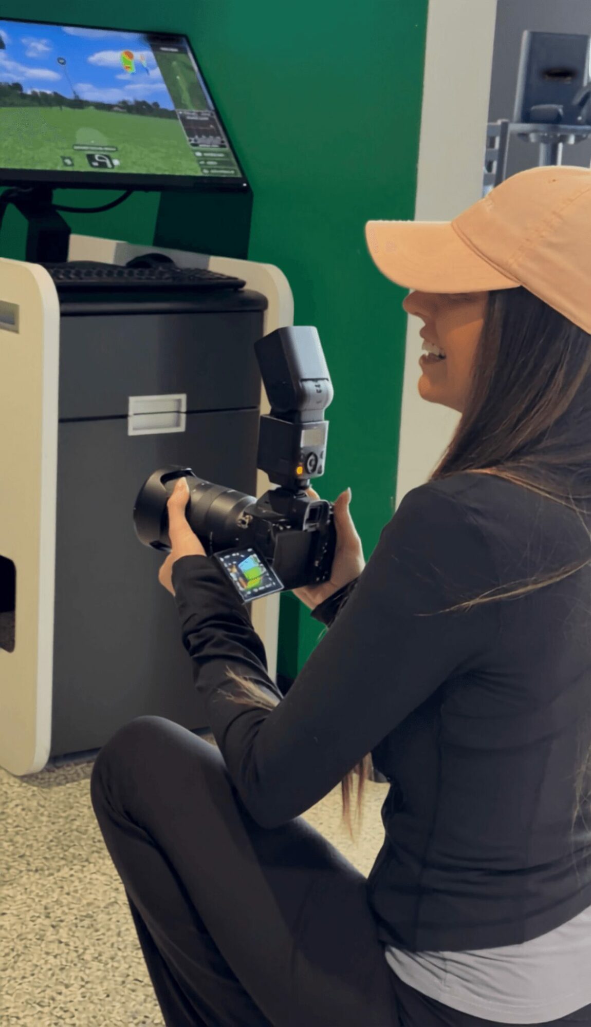 Person sitting on the floor holding a camera, wearing a beige cap and black jacket, with a computer station nearby.
