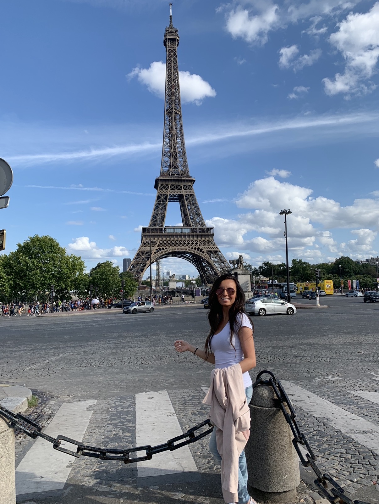 Woman standing near a chain barrier in front of the Eiffel Tower on a sunny day with blue sky and clouds.