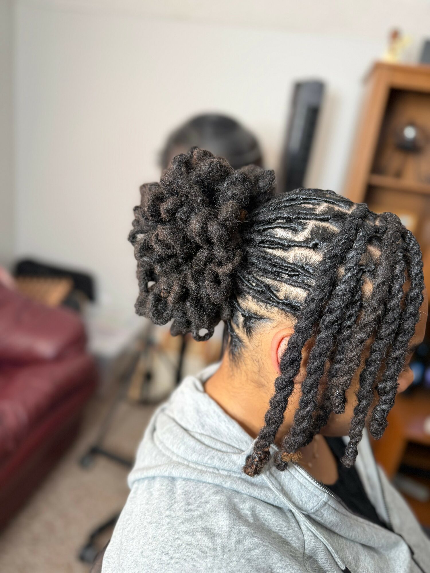 Person with styled hair, including a large curly bun and long twists, in a room with furniture and a bookshelf