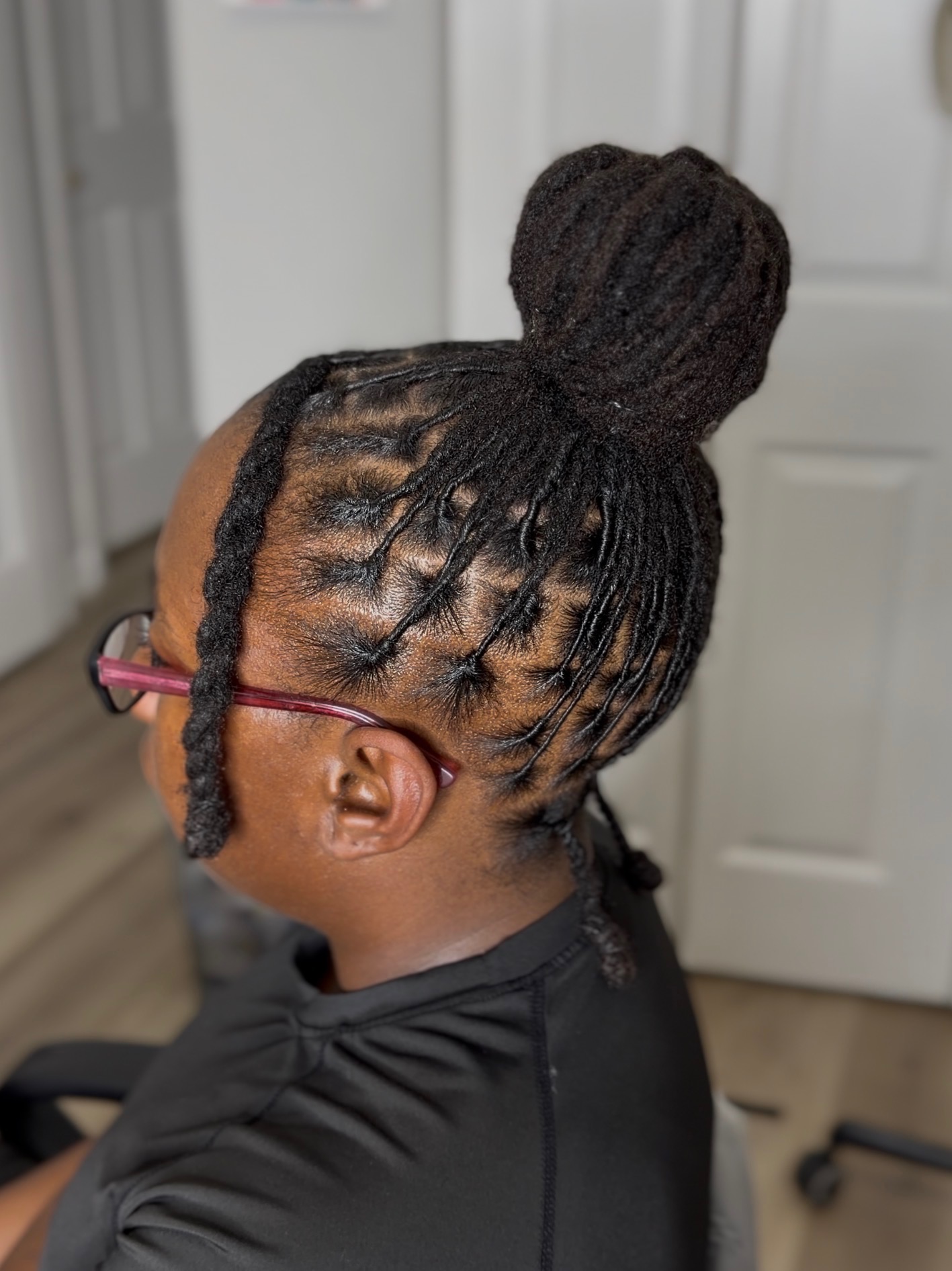 Person with braided hairstyle in a bun, wearing glasses, facing left, indoors, with a door in the background.