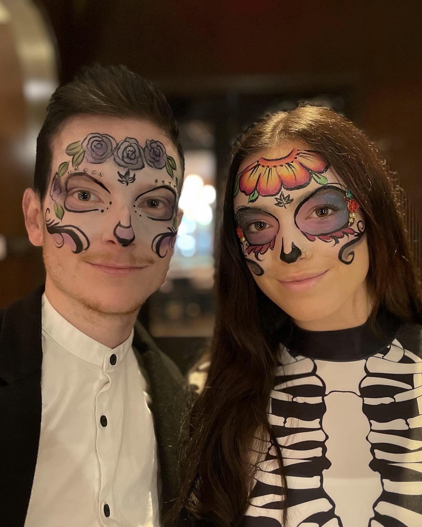 Two people with face paint, smiling, one with floral and swirl designs, the other with sugar skull and skeleton shirt.