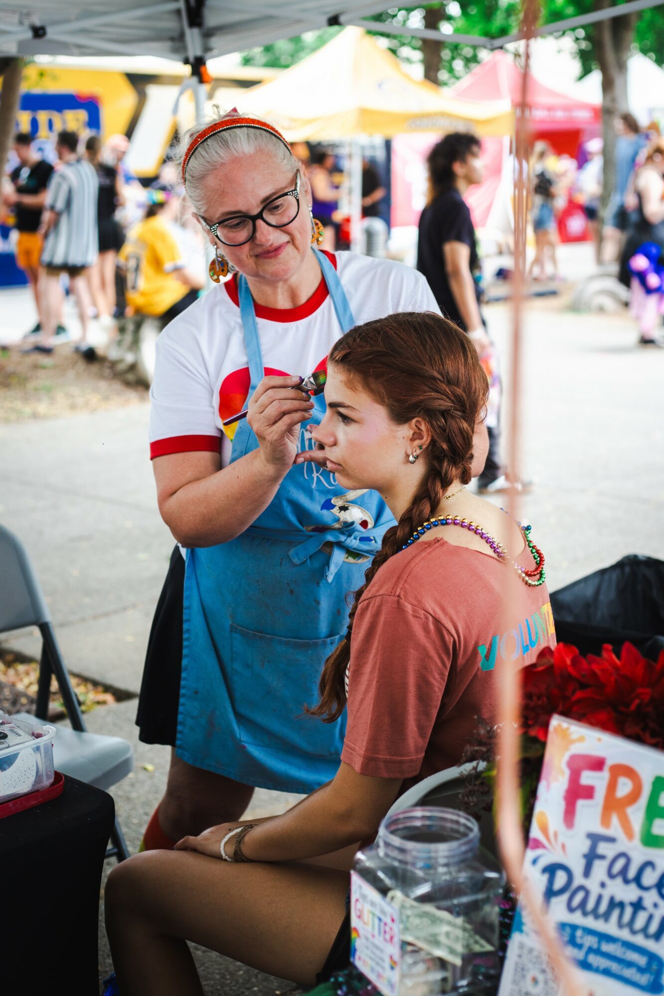 Woman painting a child's face at an outdoor event with tents and people in background.
