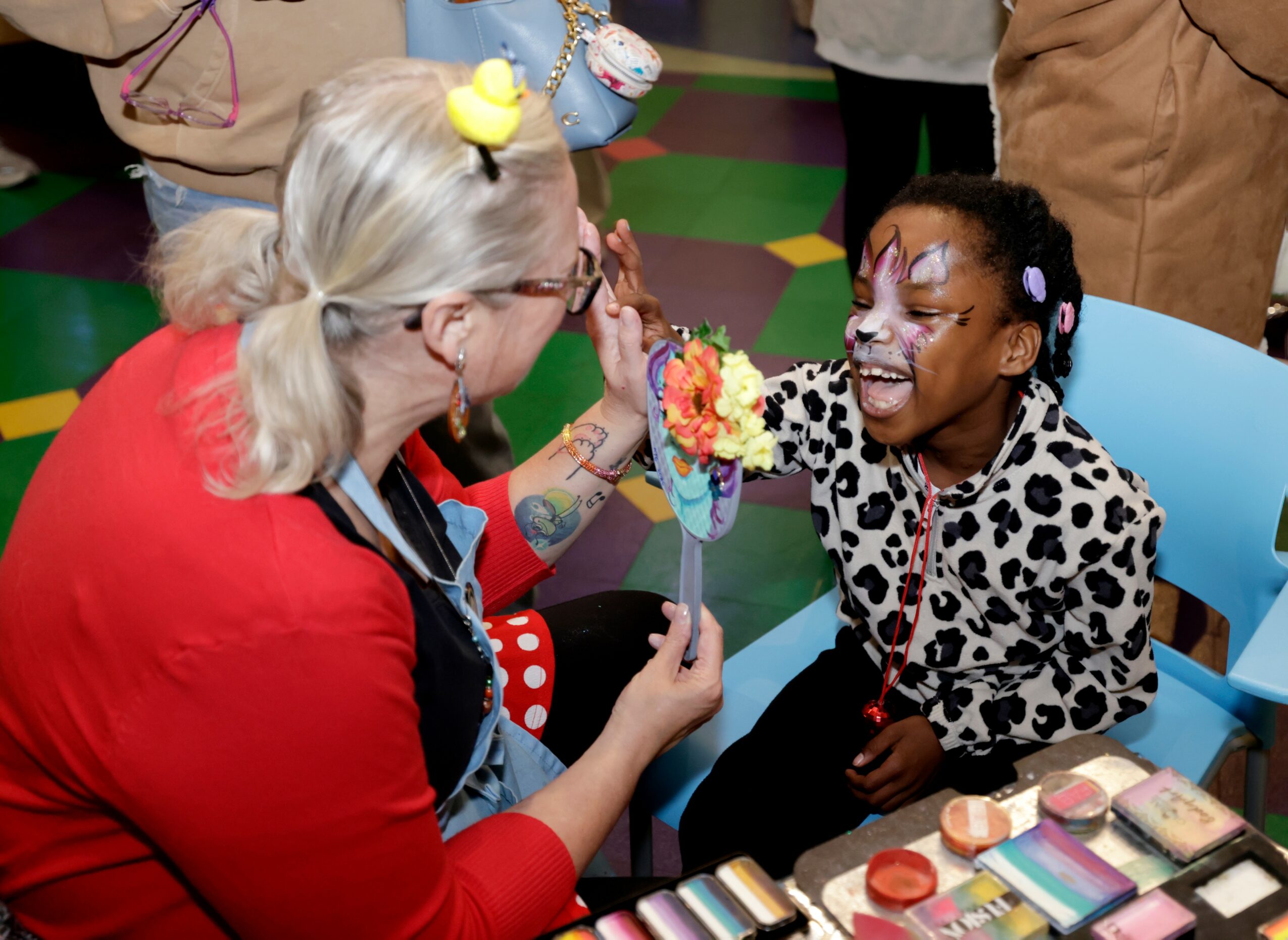 Adult woman and young girl smiling, girl with face paint, colorful makeup, and a toy on her head, sitting at a table with makeup products.