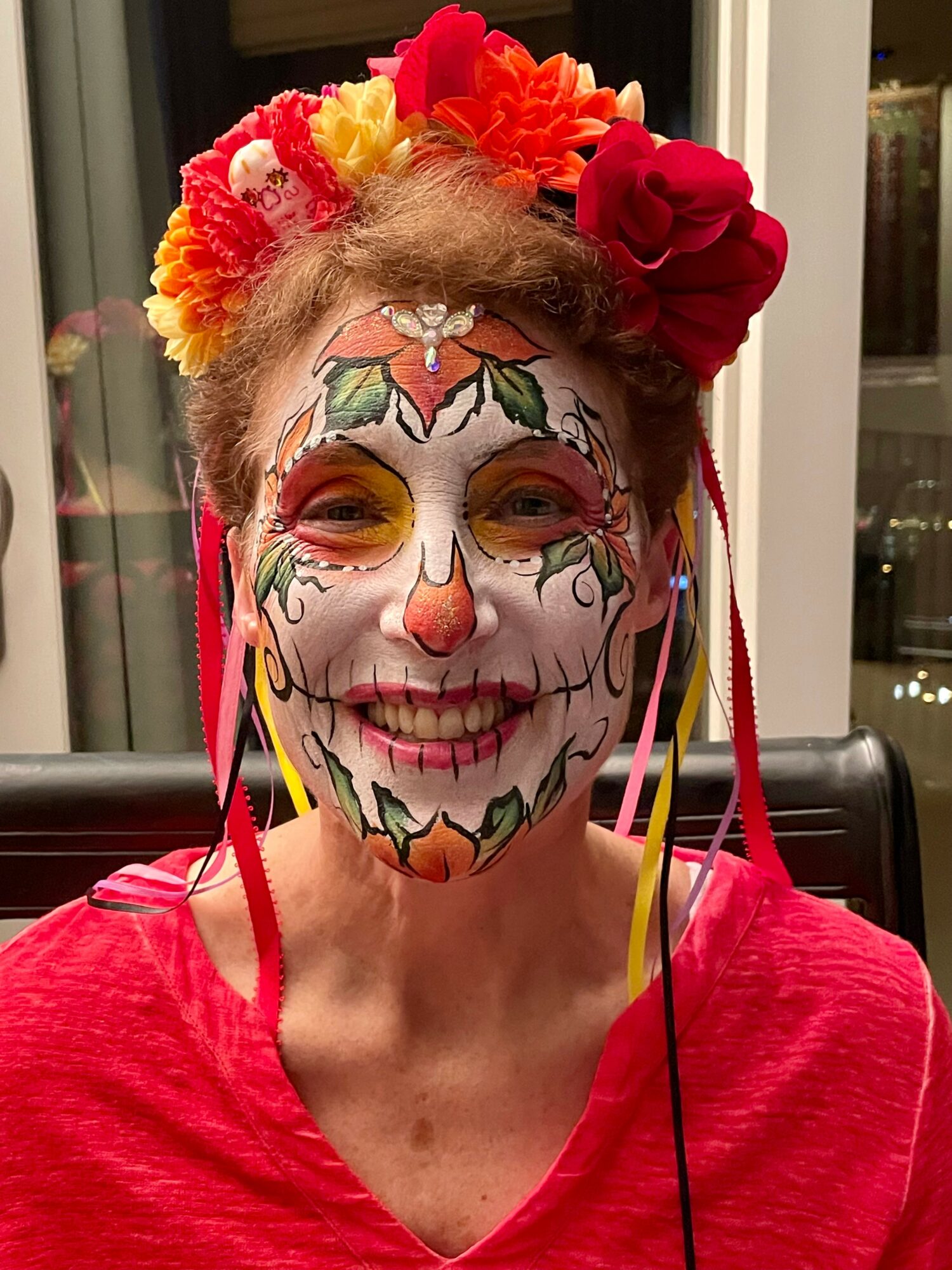 Woman with colorful face paint resembling a sugar skull, wearing a red top and floral headpiece, smiling.
