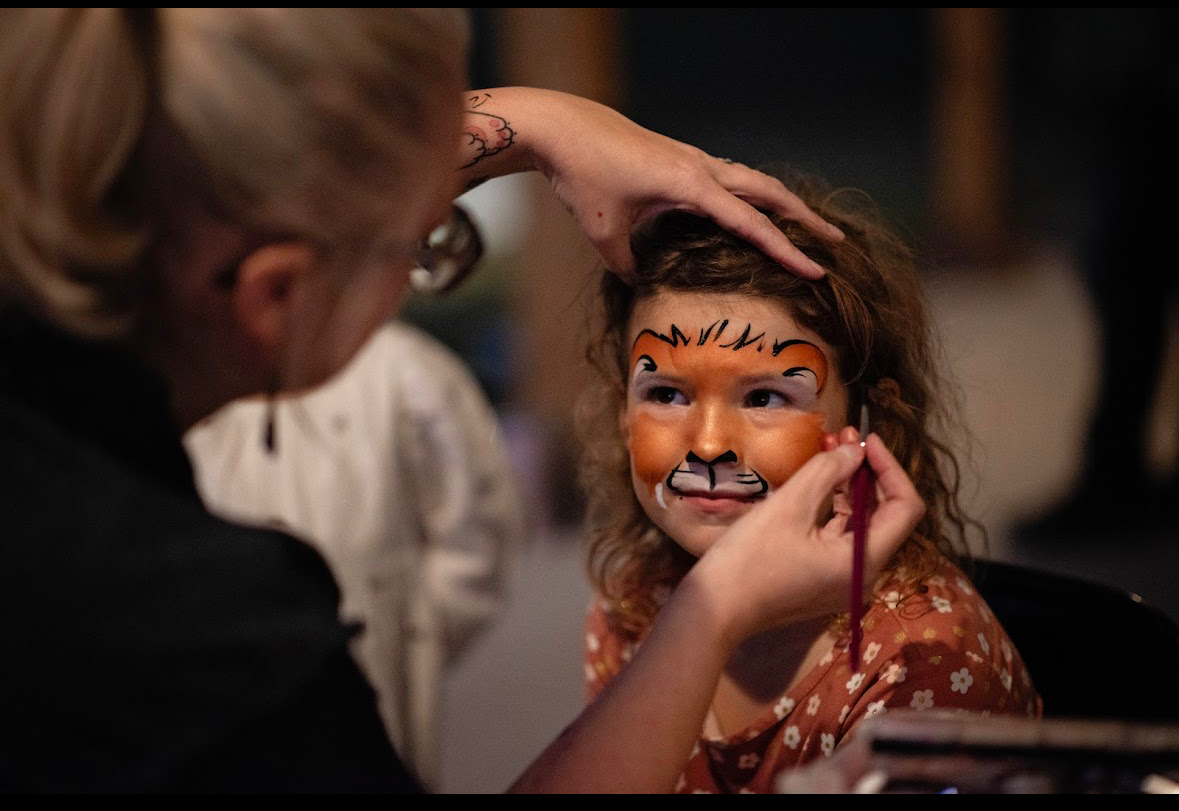 Child with face painted as a tiger, smiling, while a person paints more details on her face.