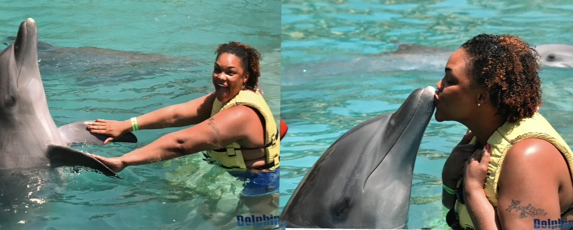 Person touching dolphin's nose in water, smiling, with another person close to dolphin's face, both in a pool.