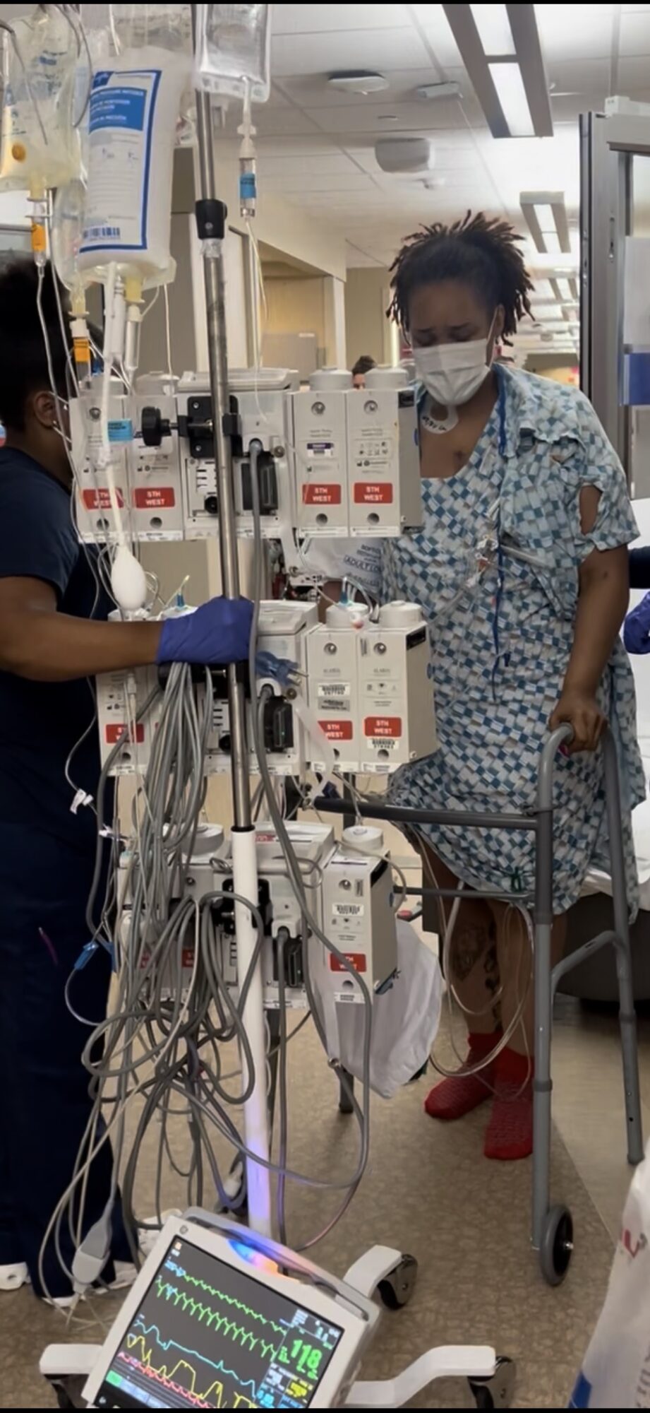 Two healthcare workers in a hospital room, one in scrubs and mask, the other in patterned gown and mask, near medical equipment.