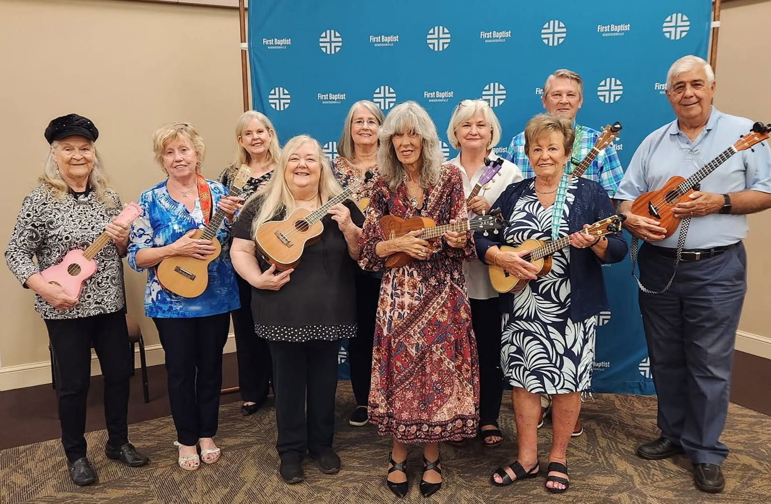 Group of ten women and one man holding ukuleles, standing indoors in front of a blue backdrop with white logos.