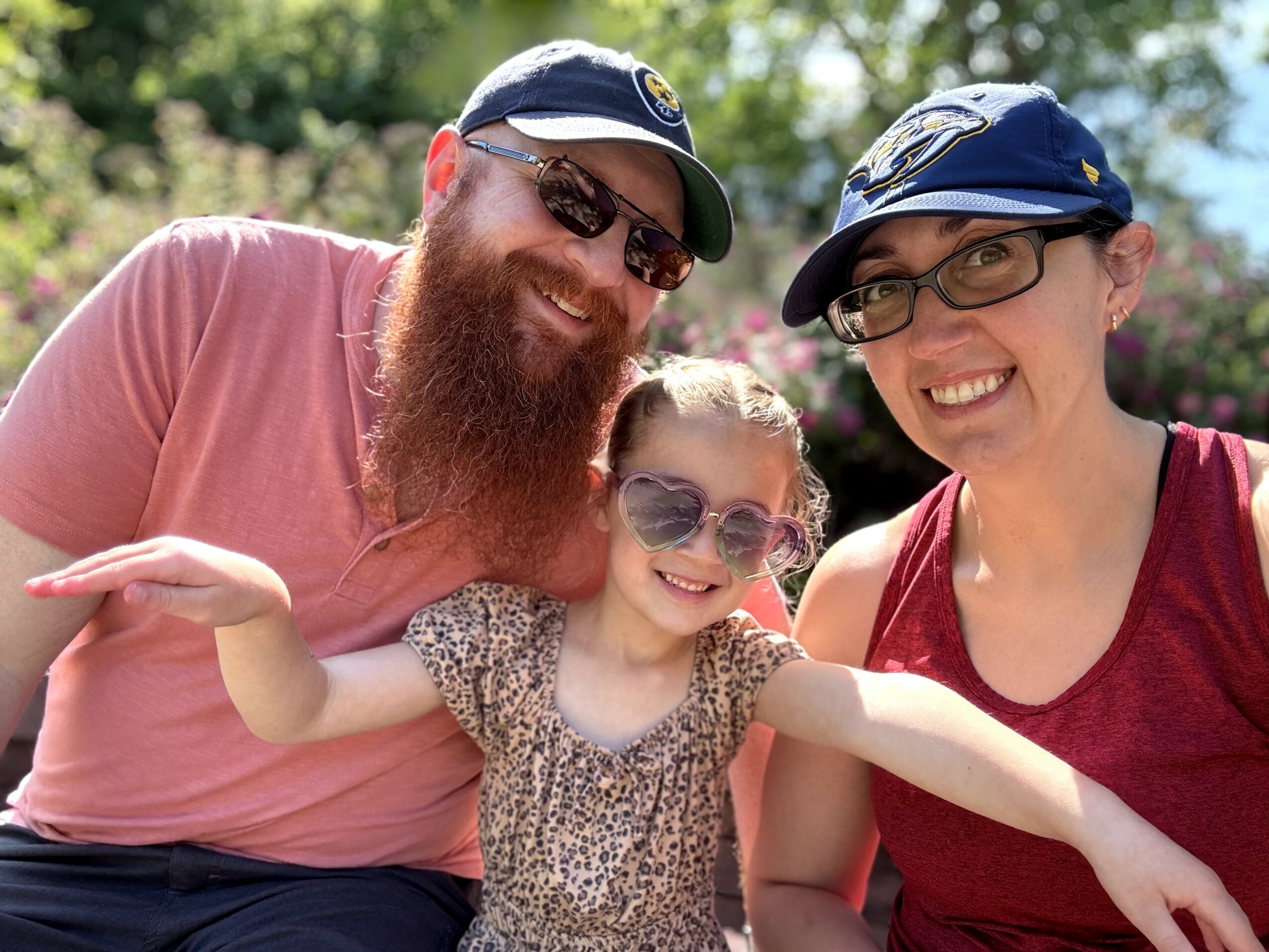 Three people smiling outdoors, two adults and a child, all wearing sunglasses and hats, with trees in the background.