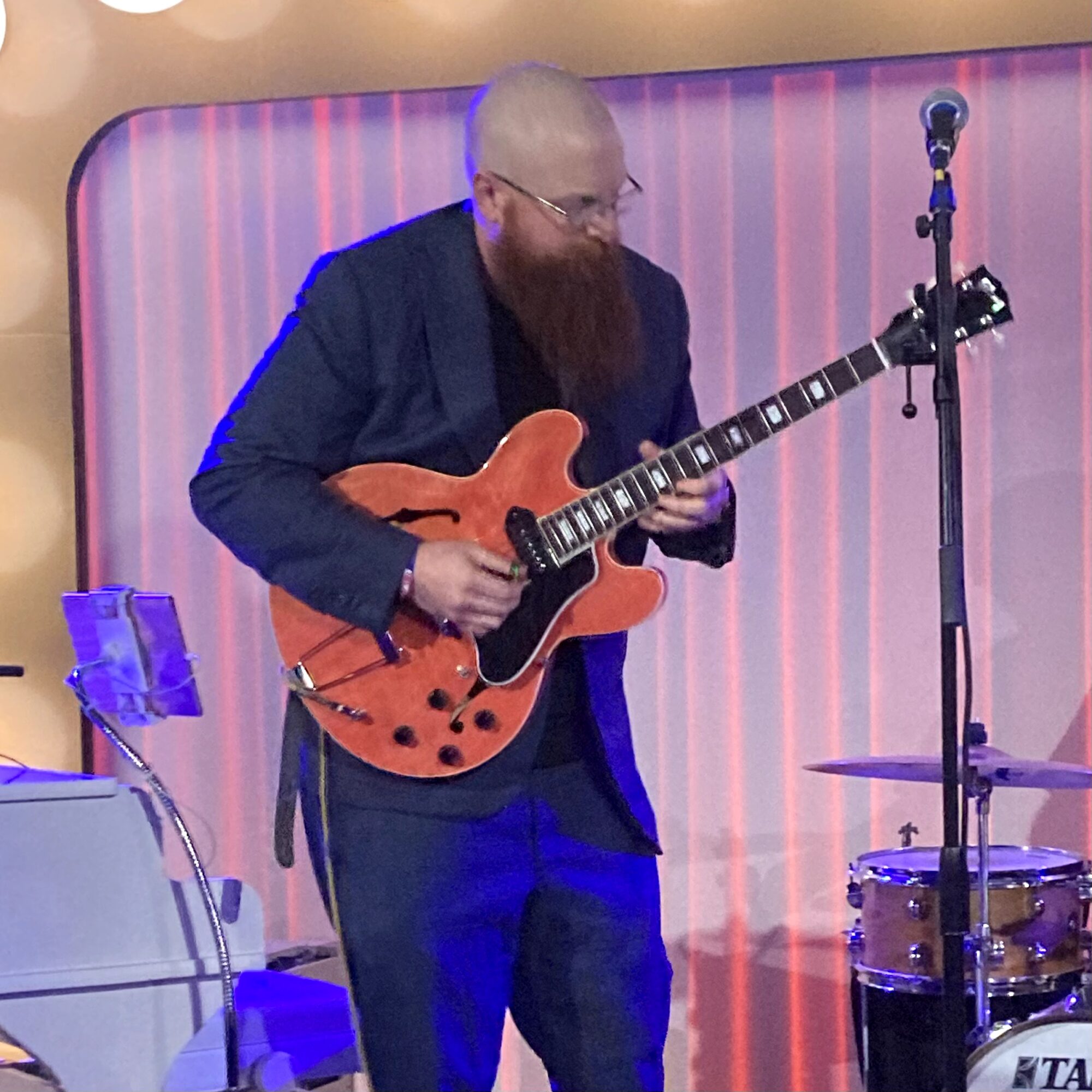 Man with a beard playing an electric guitar on stage with a microphone and drums in the background.