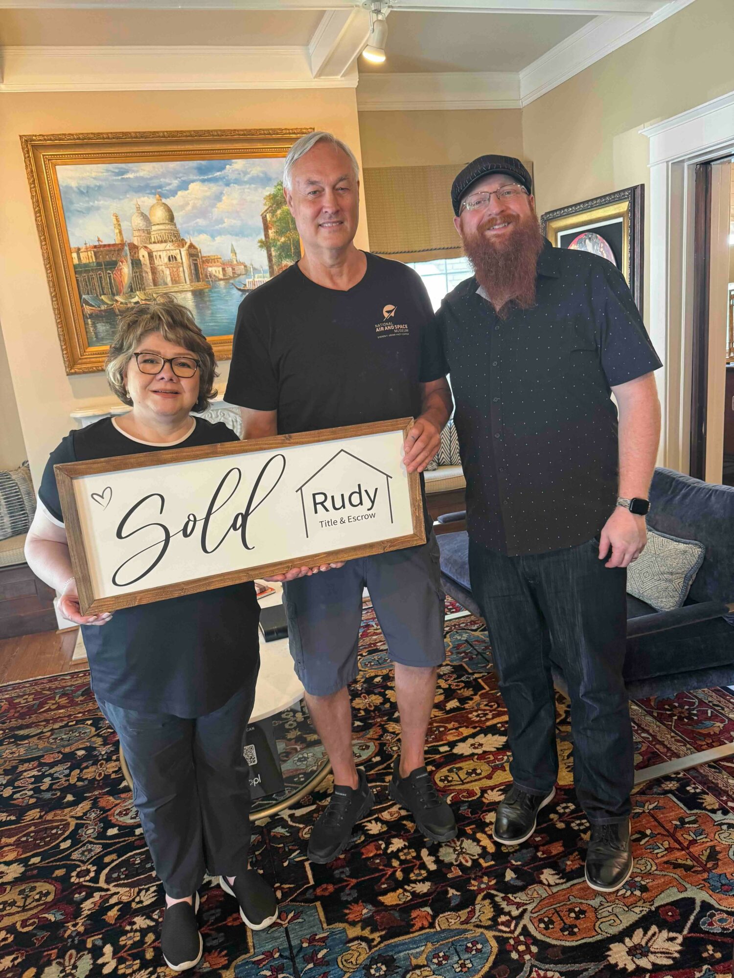 Three people standing indoors, holding a 'Sold' sign, smiling, with a painting and a sofa in the background.