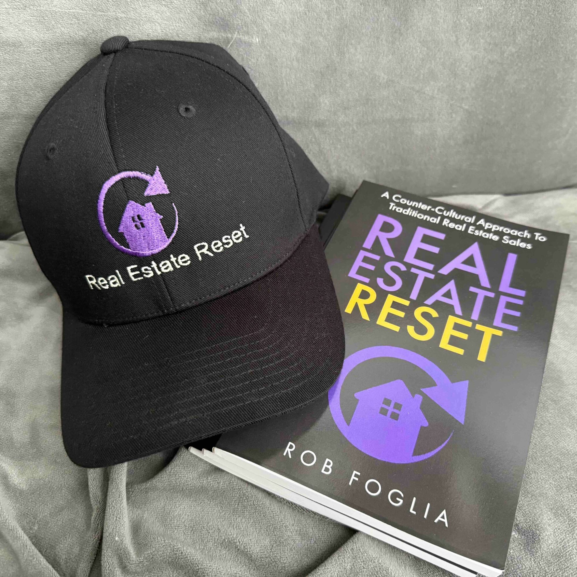 Black cap with purple logo and text, next to a book titled 'Real Estate Reset' by Rob Foglia, on gray fabric.