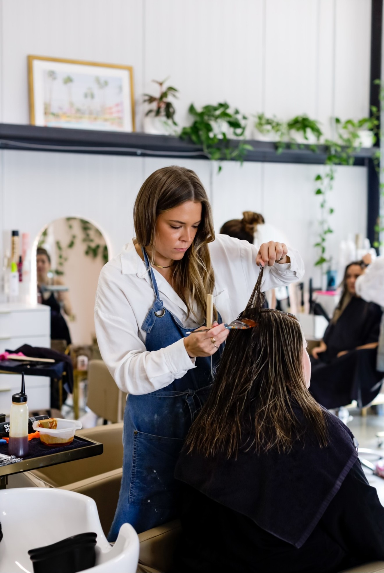 Hairdresser using scissors on a client's hair in a salon with mirrors and plants, other clients seated nearby.
