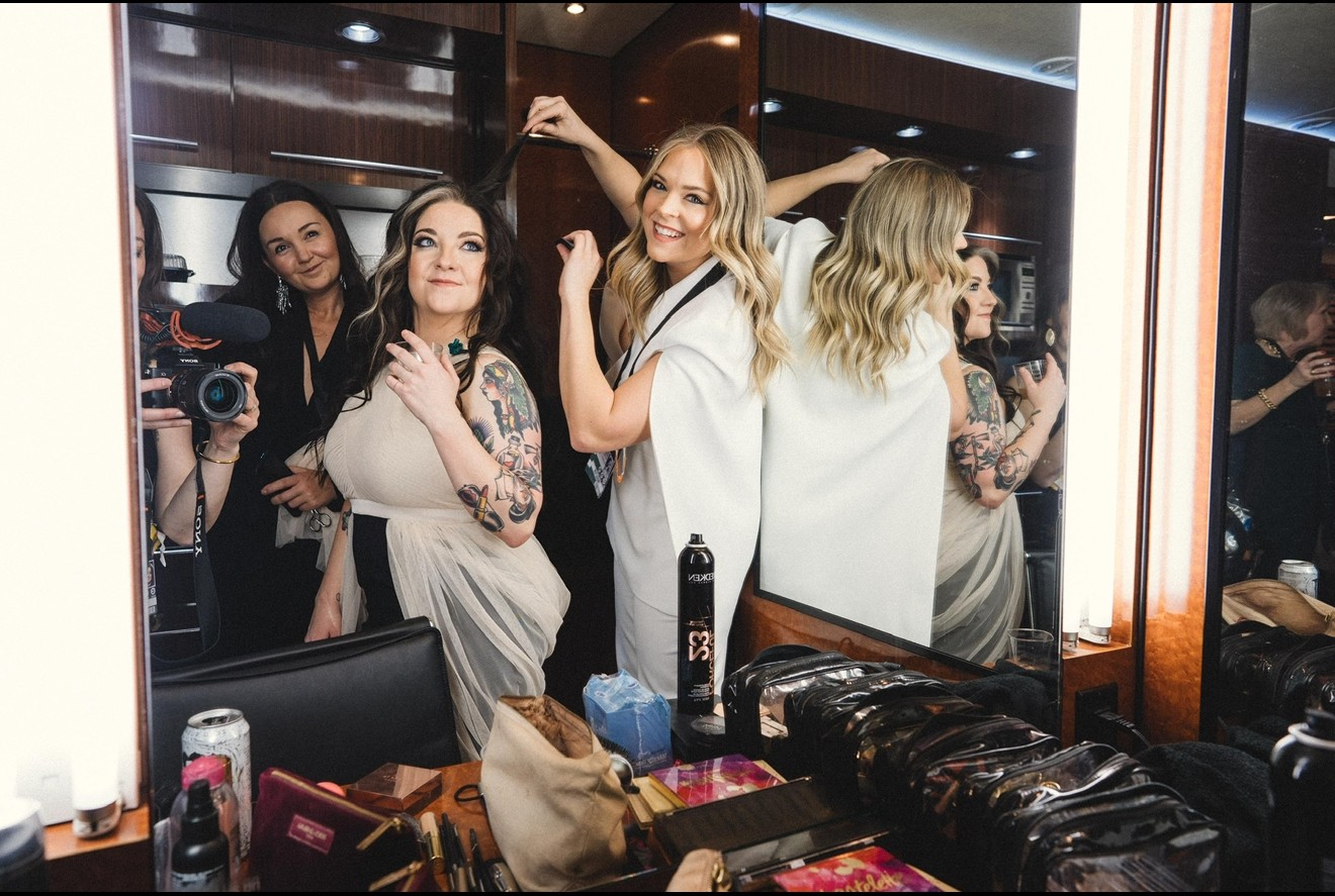 Three women in a dressing room, two with long hair, one with short hair, smiling and posing, with mirrors and makeup items visible.