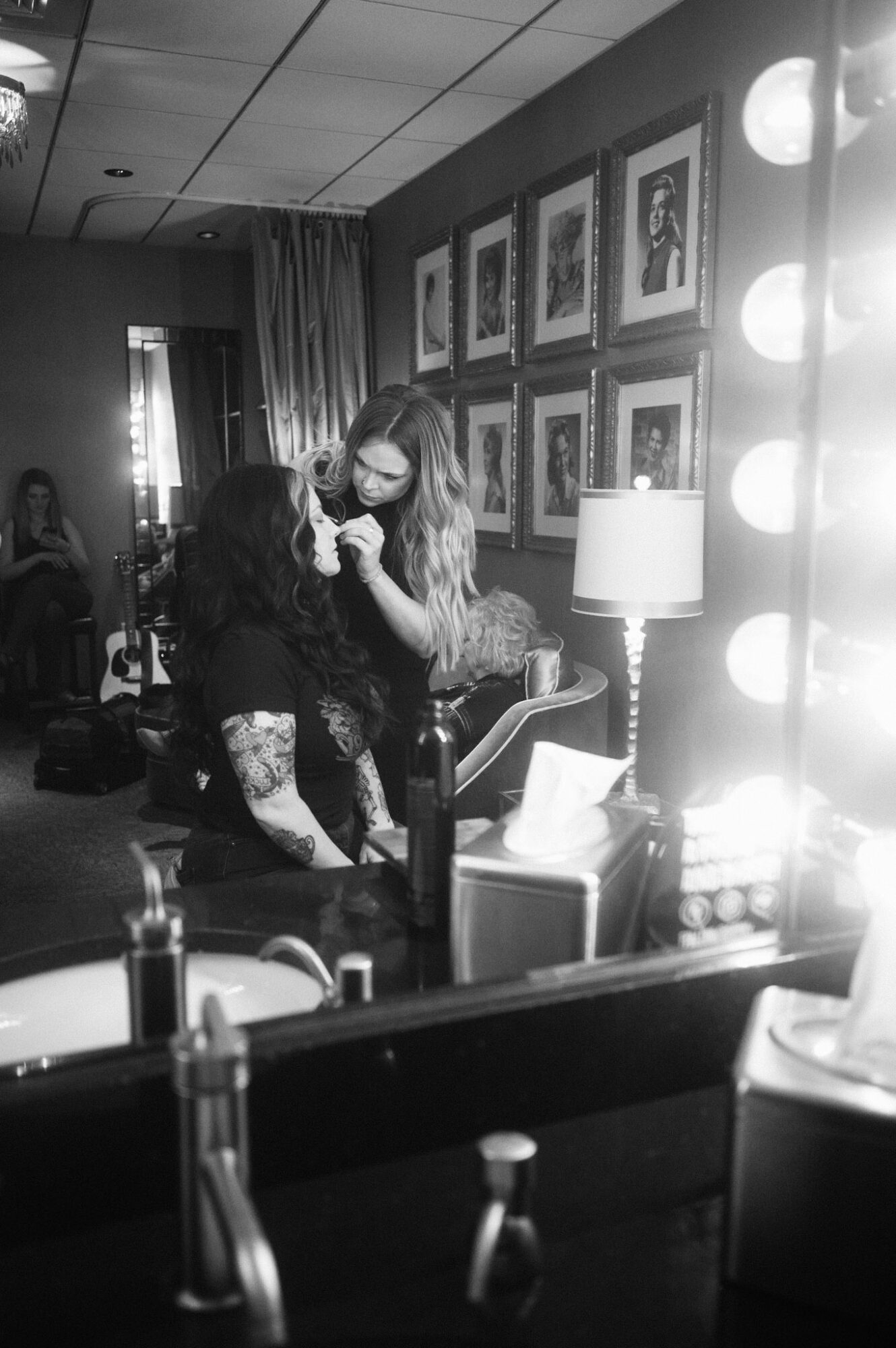 Two women in a room with framed pictures on the wall, one applying makeup to the other, with a lamp and mirror nearby.