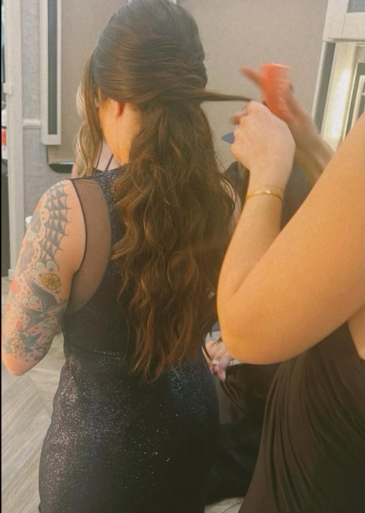 Person with long, wavy hair getting their hair styled by another person, showing a tattooed arm, in an indoor setting.