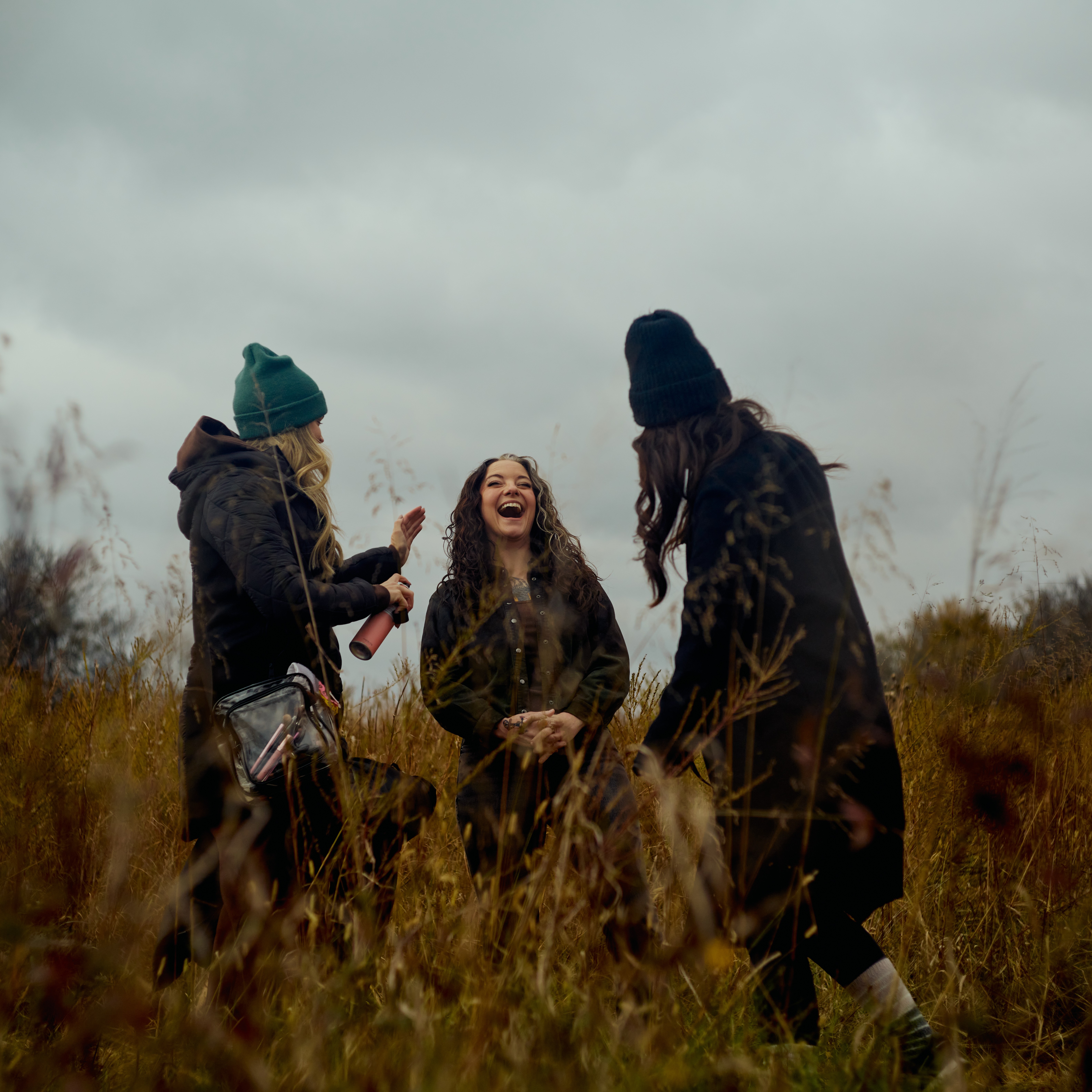 Three women standing outdoors in a grassy field, smiling and talking, with cloudy sky overhead.