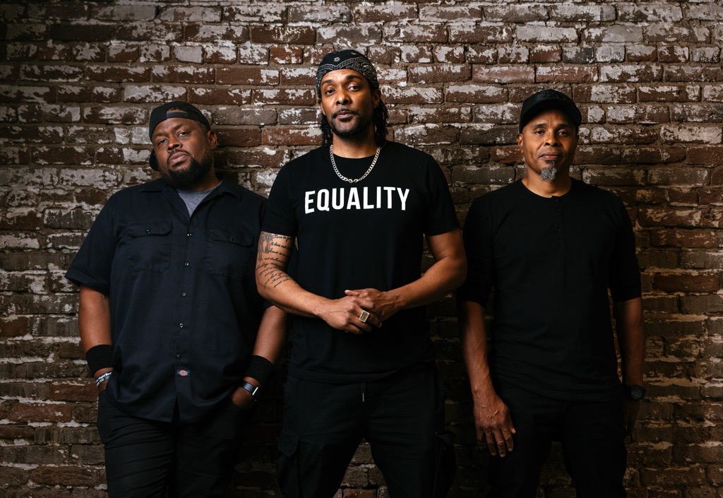 Three men standing against a brick wall, one wearing a shirt with 'EQUALITY' written on it.