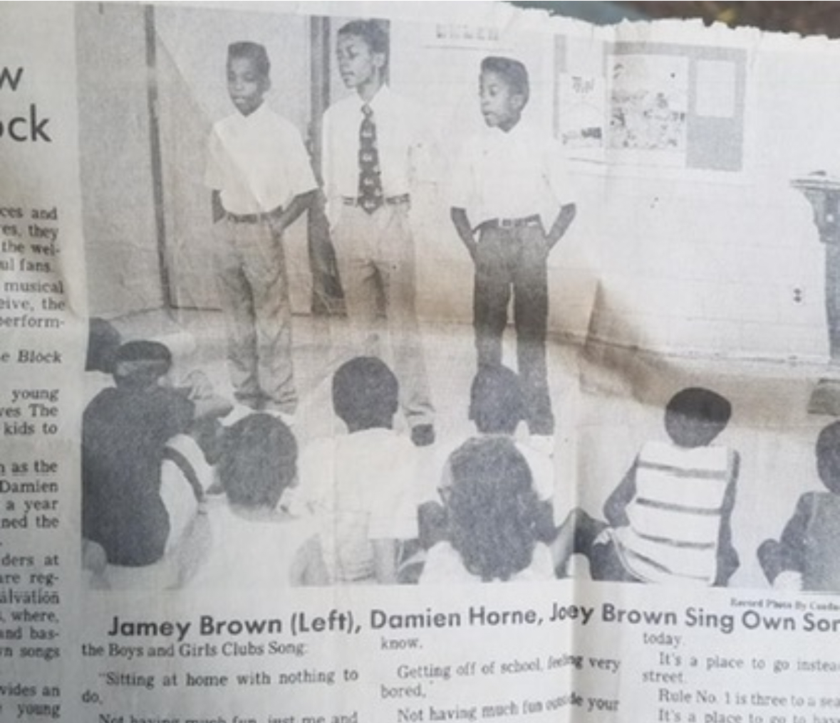 Three children stand in front of a classroom, with several children seated on the floor facing them, in a black-and-white photo.
