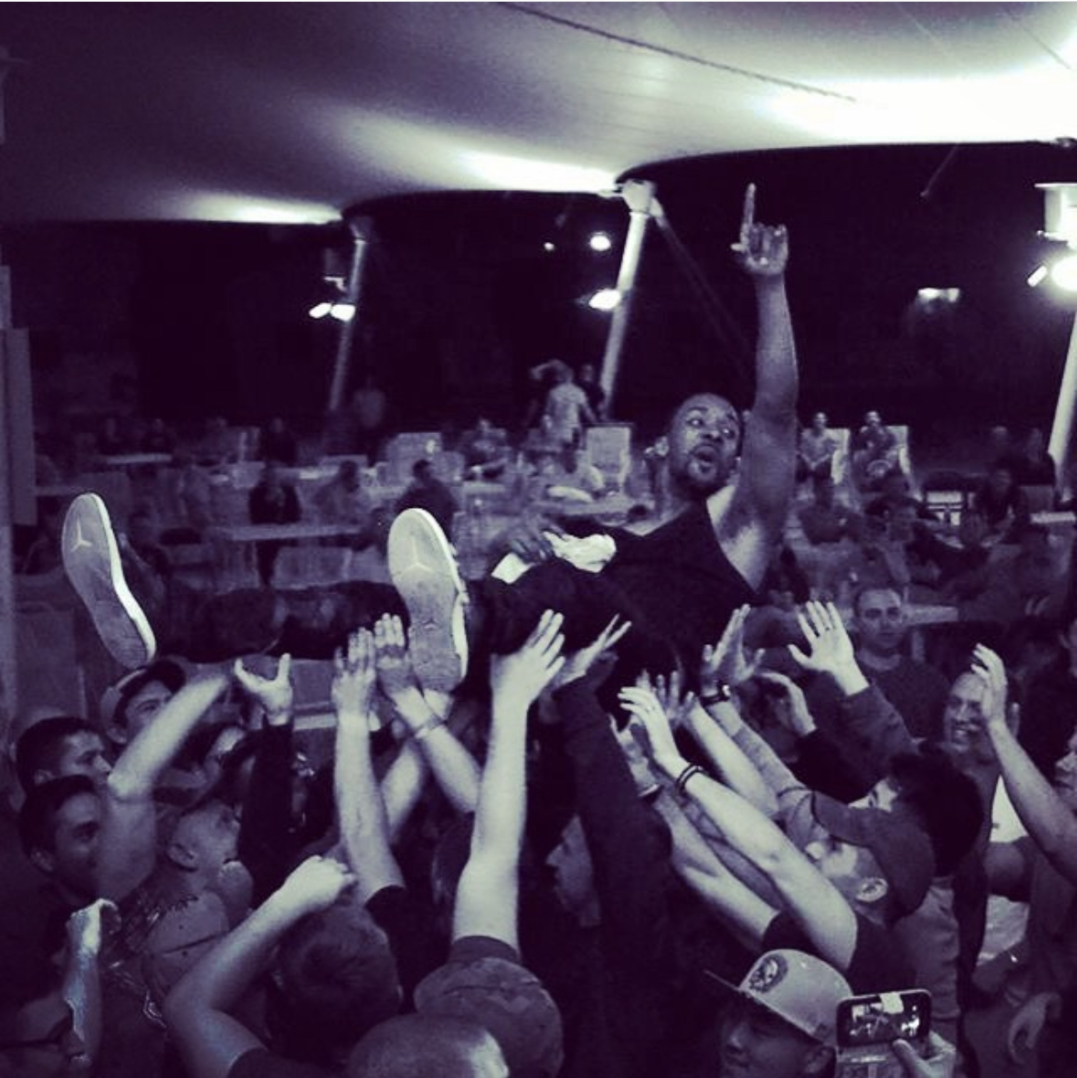 Person crowd surfing at a concert, raised above hands of audience, with people in background under a tent-like structure.