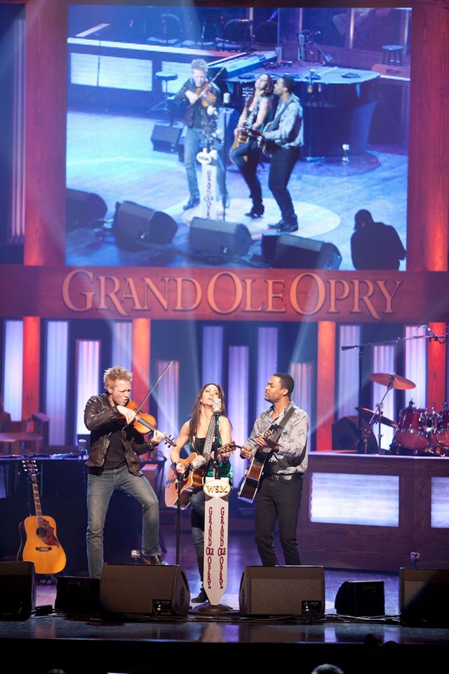 Three musicians perform on stage with a large screen above showing a close-up of them, in front of a red curtain and blue backdrop.