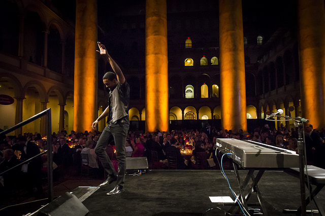 Performer dancing on stage with audience in a large, illuminated indoor venue with tall columns and multiple levels.