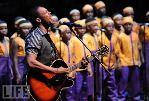 Man playing guitar and singing in front of a choir of children wearing yellow and purple uniforms on stage.