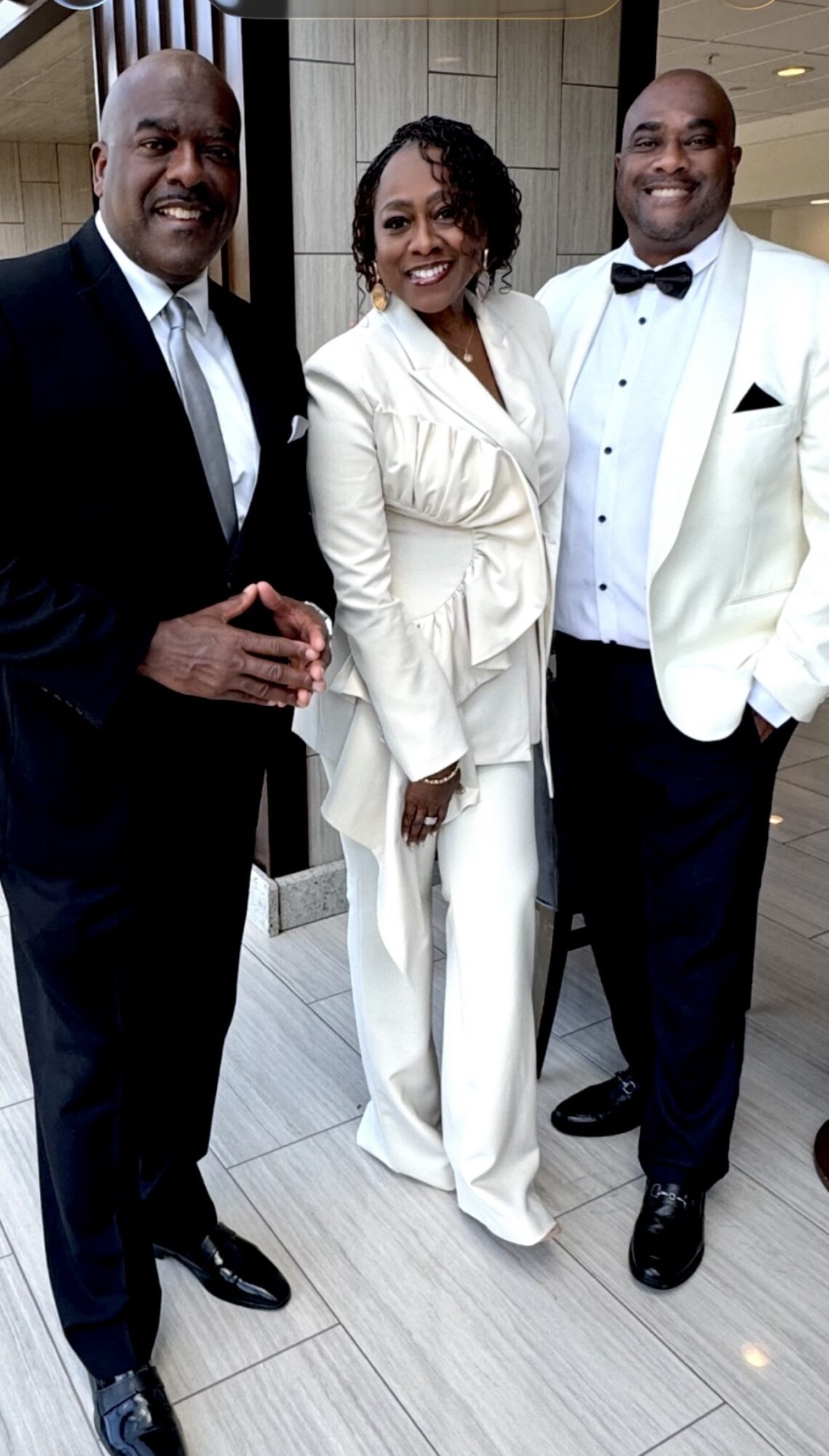 Three people dressed in formal attire standing together indoors, smiling at the camera.