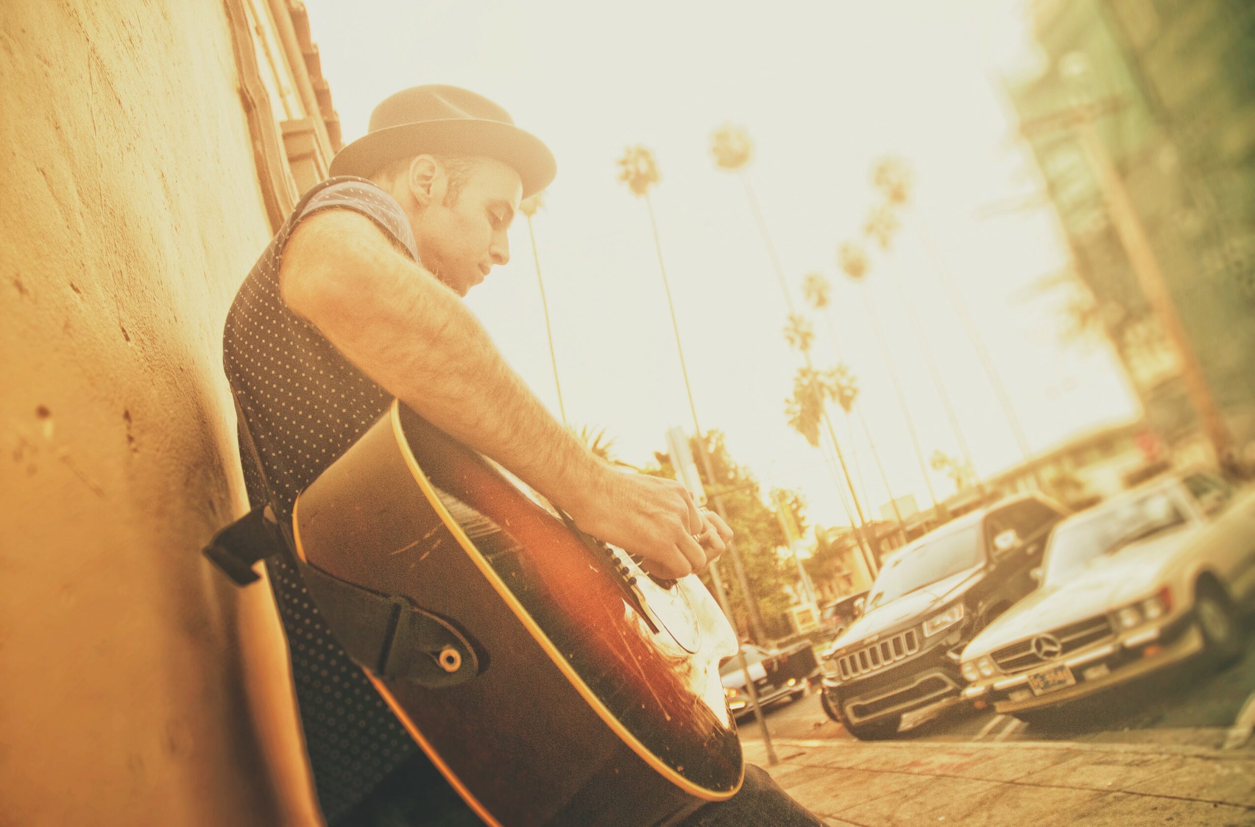 Person playing guitar outdoors with parked cars and palm trees in background, sunlight brightening scene.