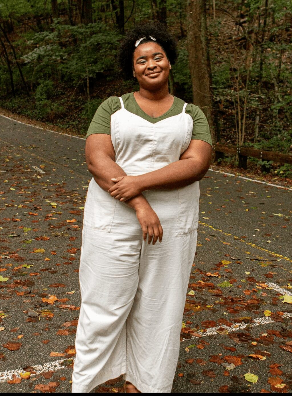 Person standing outdoors on a leaf-covered path, smiling, wearing white overalls and green shirt, surrounded by trees.