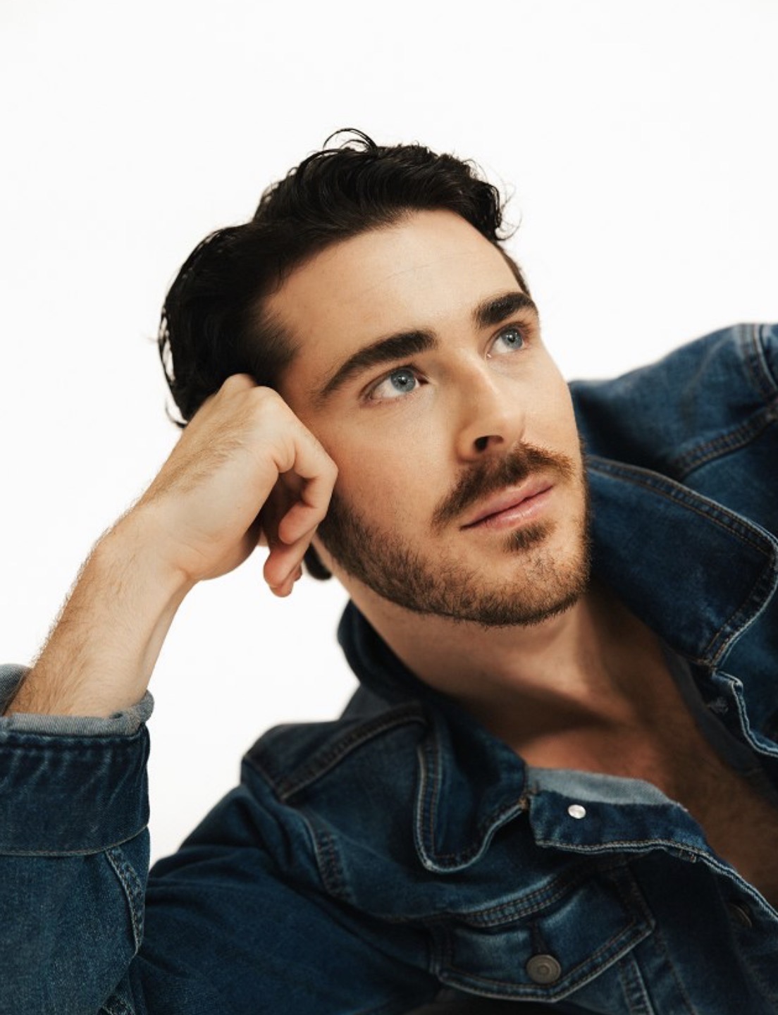 Young man with dark hair and beard, lying down with head resting on hand, looking upward, wearing a denim jacket.