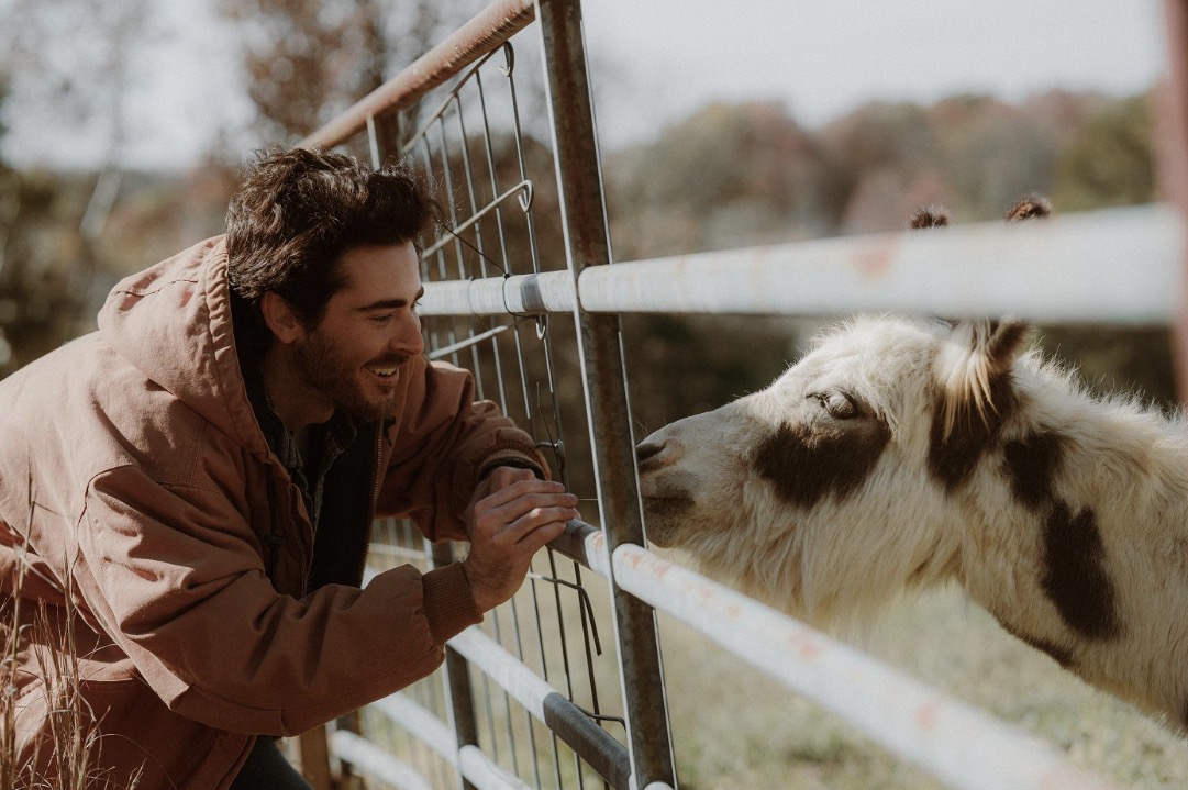 Person smiling and leaning on a fence, interacting with a goat on the other side outdoors.