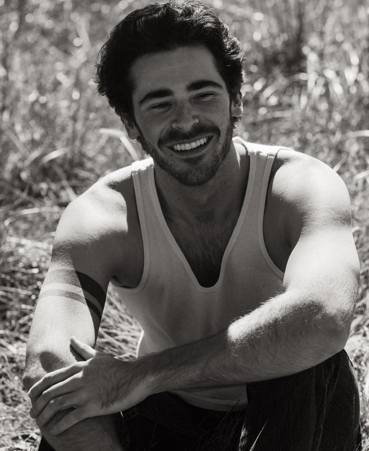 Young man with dark hair and beard smiling, wearing a sleeveless shirt outdoors, arms crossed, in natural setting