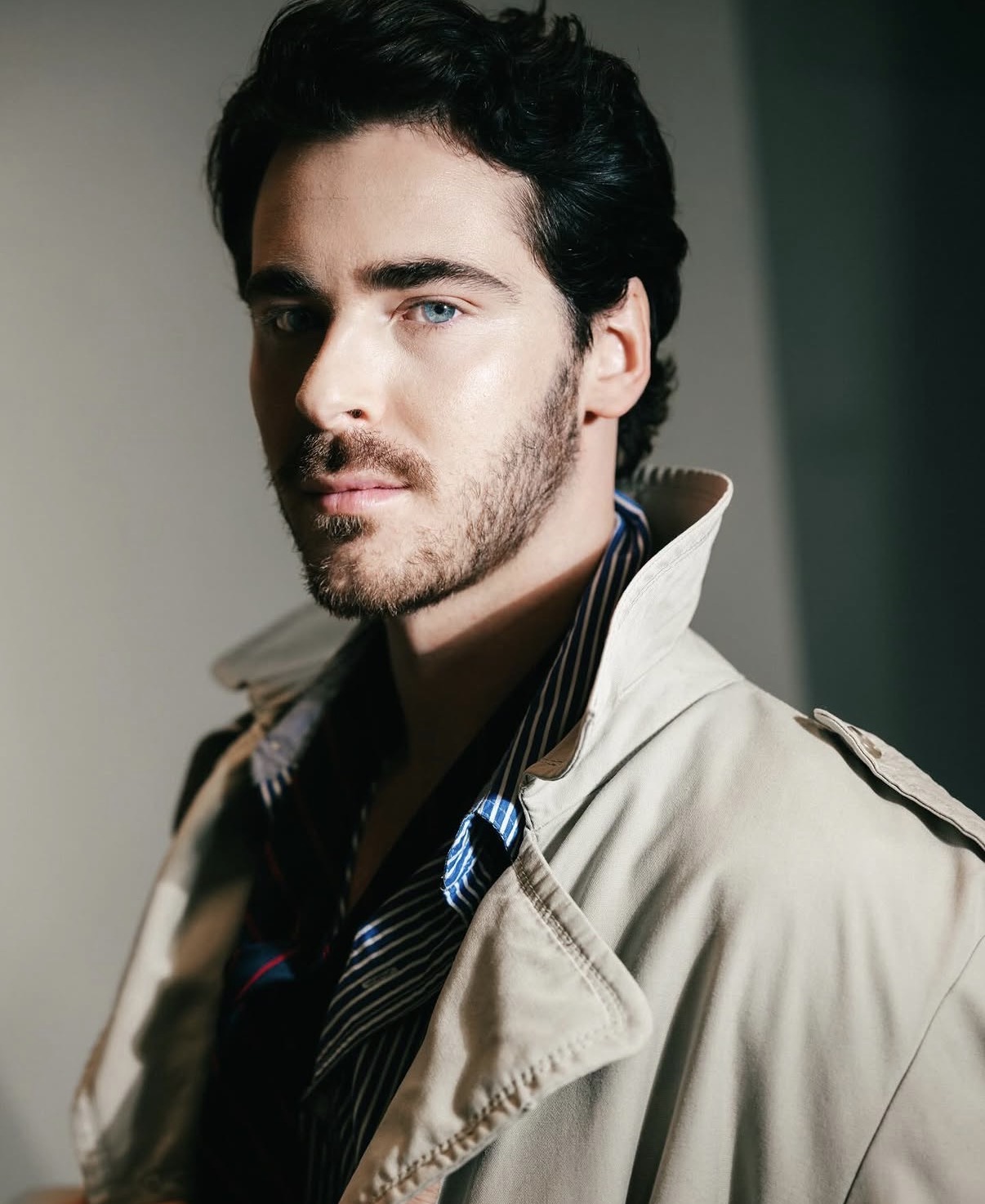 Portrait of a man with dark hair and beard, wearing a beige trench coat and black shirt, looking at the camera.