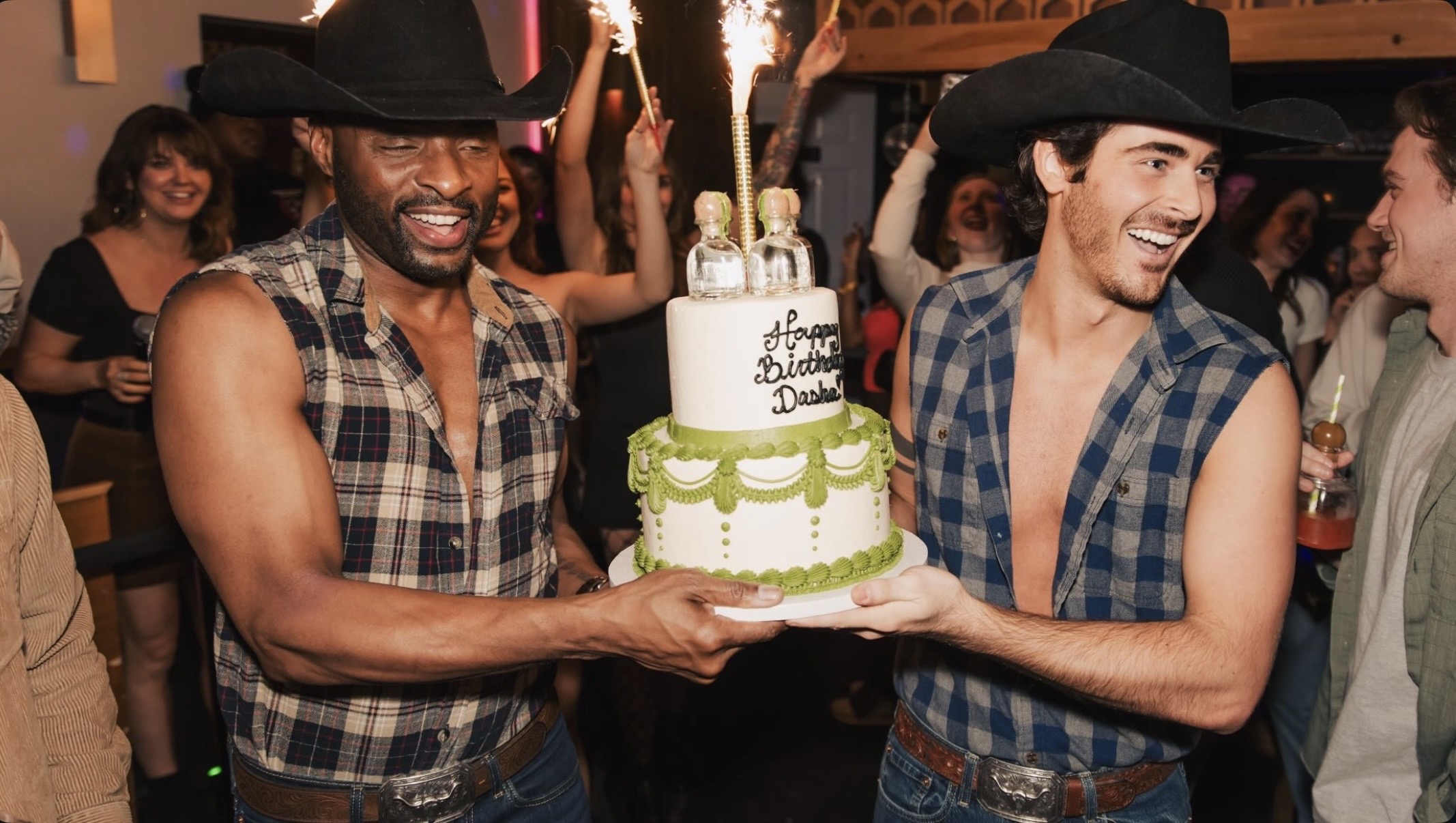 Two men in plaid shirts and cowboy hats hold a birthday cake with candles, smiling at a celebration.