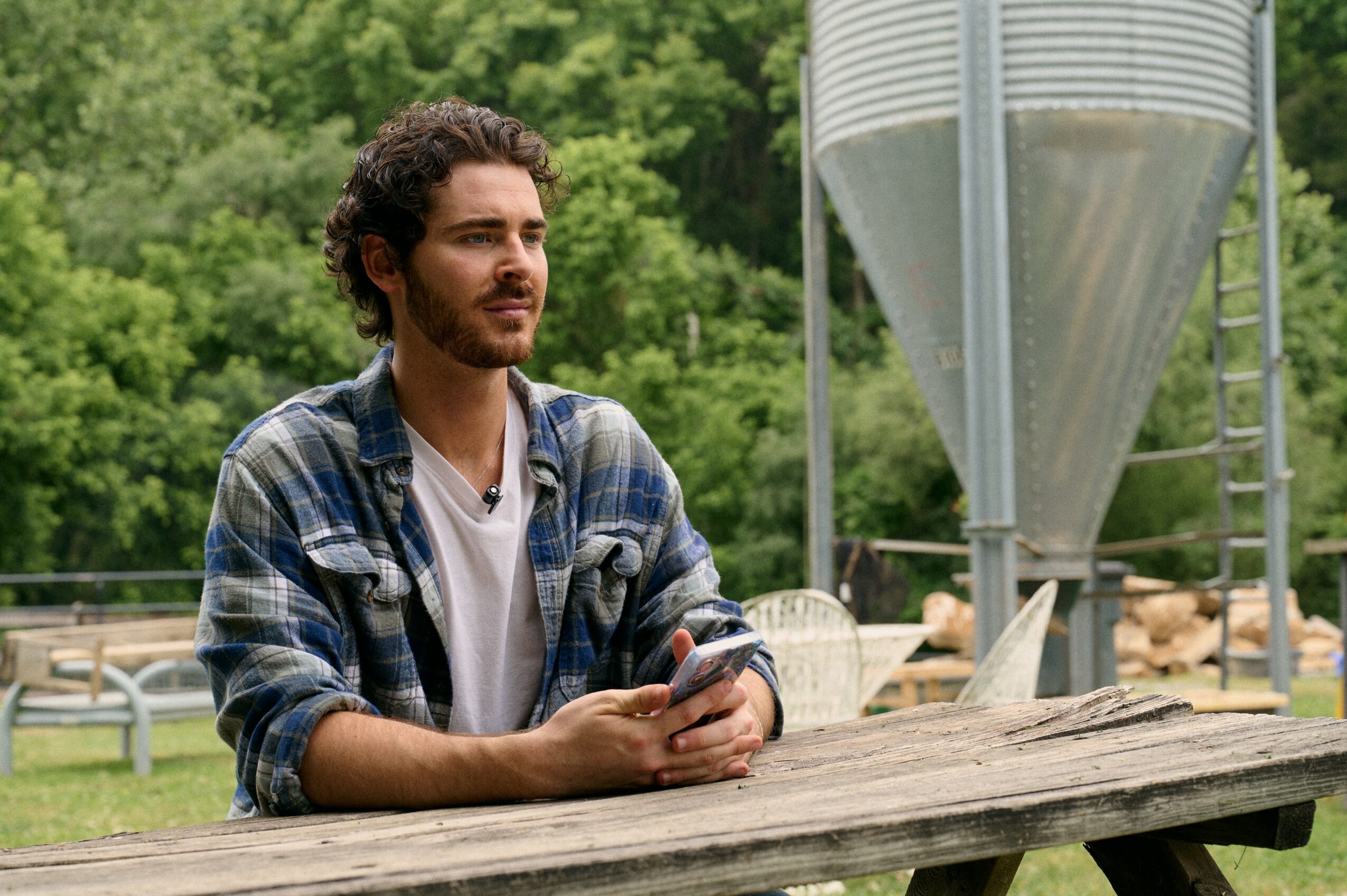 Man with curly hair and beard sitting at a wooden table outdoors holding a phone, with a large silo in the background.